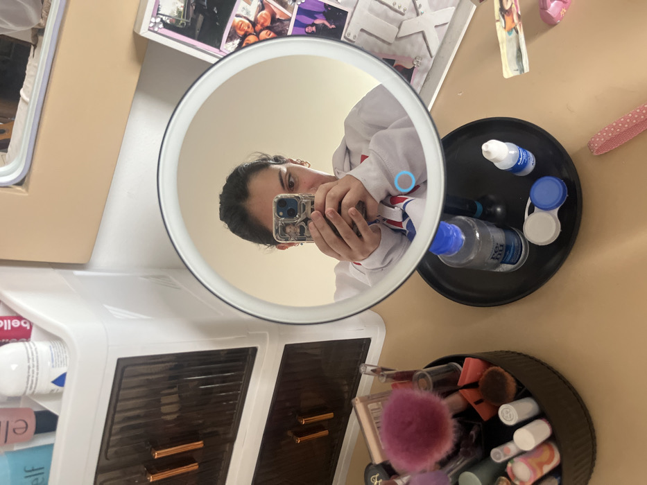 A vanity setup is captured through a circular mirror. Reflected in the mirror is a person holding a phone. A black tray holding a bottle of contact lens solution, eye drops, and a contact lens case sits beside the mirror. Below the tray is a container overflowing with makeup brushes and cosmetics. A white storage unit with brown drawers and shelves filled with toiletries is also visible. A picture frame and other miscellaneous items are scattered on the surrounding surface.