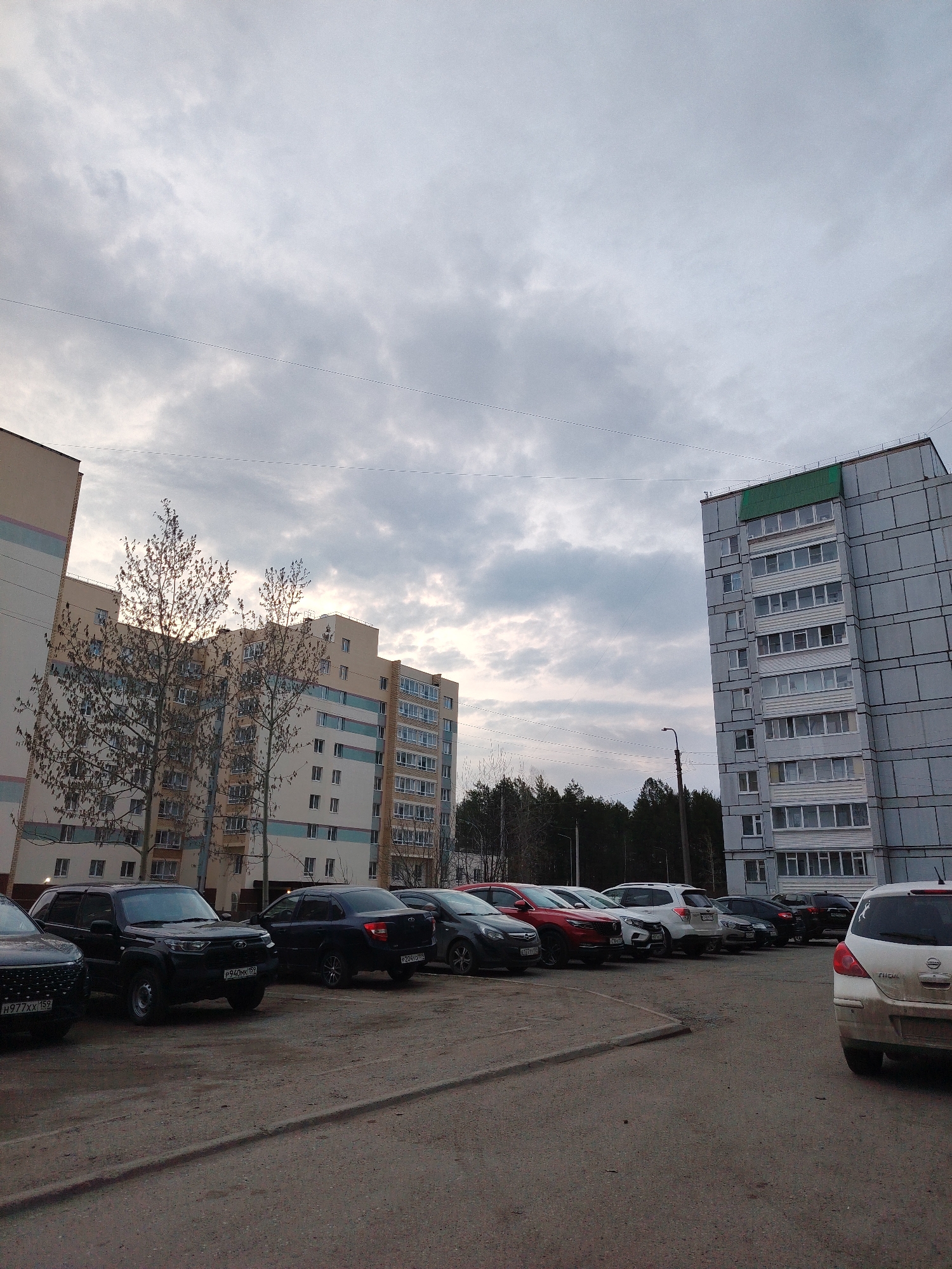 The image depicts a parking lot in an urban environment, likely in Unknown, Unknown. Several cars of various makes and models are parked in rows. In the background, there are multi-story residential buildings with light-colored facades and a building with a gray façade. The sky is overcast with clouds. Bare trees are visible near the buildings. A street lamp is present near the gray building. The ground is paved and shows signs of wear and tear.