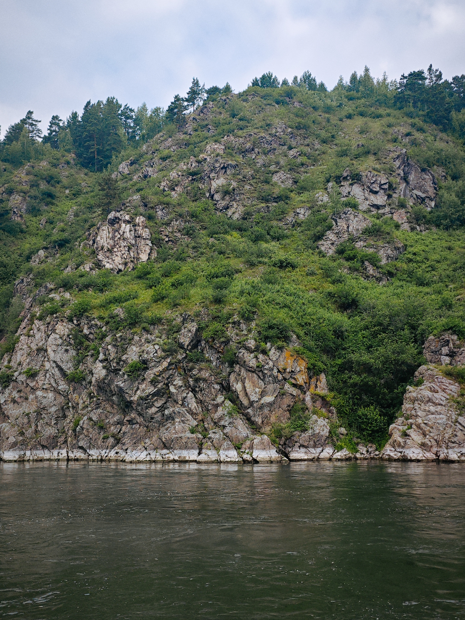 The image depicts a rocky cliff face overgrown with dense green foliage and scattered trees, rising from the dark, rippling surface of a wide river. The cliff is composed of rough, textured rock in shades of gray, brown, and white, with some patches of orange lichen. The vegetation is lush, with a variety of shrubs and trees forming a thick canopy along the slopes. The sky above is overcast with a pale gray, cloudy appearance. The scene appears to be a natural landscape, possibly a riverbank or a mountainside overlooking a body of water. The location is identified as the city of Атаманово, Russia. There are no visible people or human activity in the image.