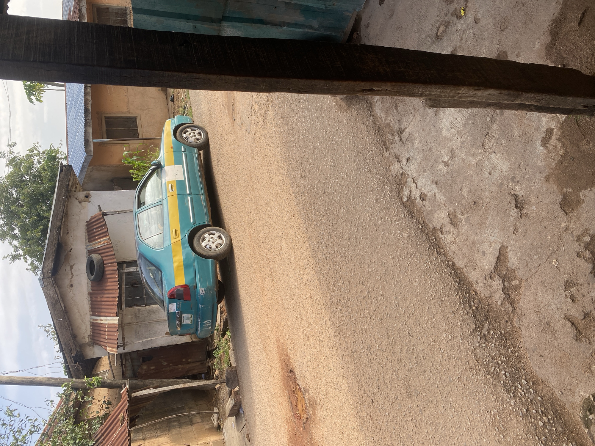 A turquoise and yellow sedan is parked on a dusty, unpaved road in front of a dilapidated building in Jos, Nigeria. The car, which appears to be a taxi, has a yellow stripe running along its side and a white square sticker on the passenger door. The building behind the car has a rusty corrugated metal roof and a tire mounted on its facade. The road surface is uneven and shows signs of wear and tear, with a small puddle of water in a pothole. The surrounding environment appears to be an informal settlement, with other structures and vegetation visible in the background. The overall scene conveys a sense of rural or semi-urban living in Nigeria.