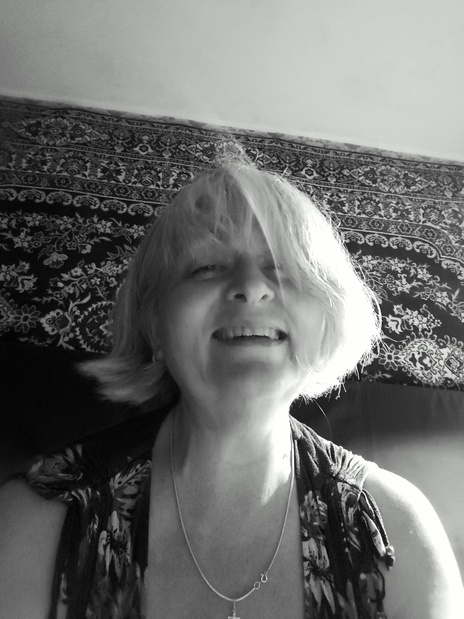A black and white, low-angle, medium close-up selfie of a middle-aged Caucasian woman with blonde hair and fair skin. She is smiling with her mouth open, showing teeth. She is wearing a dark-colored top with a floral pattern and a silver necklace with a pendant. Behind her is a dark-colored tapestry with an ornate pattern. The environment appears to be indoors, possibly a bedroom or living room, given the tapestry and likely furniture. No specific location within Krasnodar, Russia, can be identified.