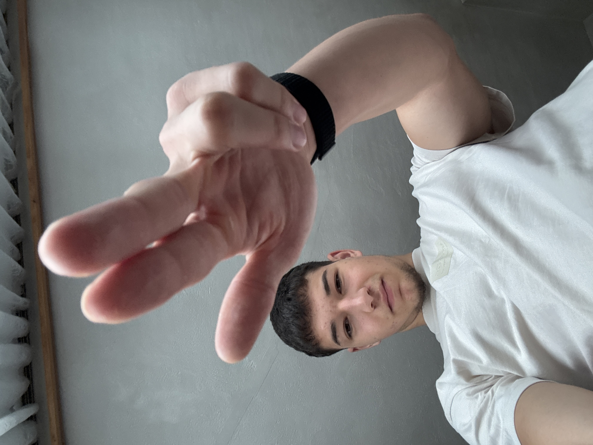 A low-angle, vertical portrait captures a young man indoors in Zlatograd, Bulgaria, against a plain light grey wall. The man, with short dark hair and light stubble, looks directly towards the camera with a subtle, relaxed smile. He is wearing a light-colored, short-sleeved t-shirt, which has a small, faint, and illegible logo or tag near the right shoulder.
His left hand is prominently featured in the foreground, extended towards the camera, with the index and middle fingers raised in a "peace" or "victory" sign. A black wristband is visible on his left wrist. The unusual perspective, looking up at the subject, suggests a casual pose or a camera placed below the man, creating a dynamic feel for the photograph. In the background on the far left, a wooden frame and what appear to be white window blinds or sheer curtains are partially visible, indicating a domestic setting. The lighting is bright and even, characteristic of daytime indoors. The overall mood of the image is casual and friendly, depicting a simple, personal moment.