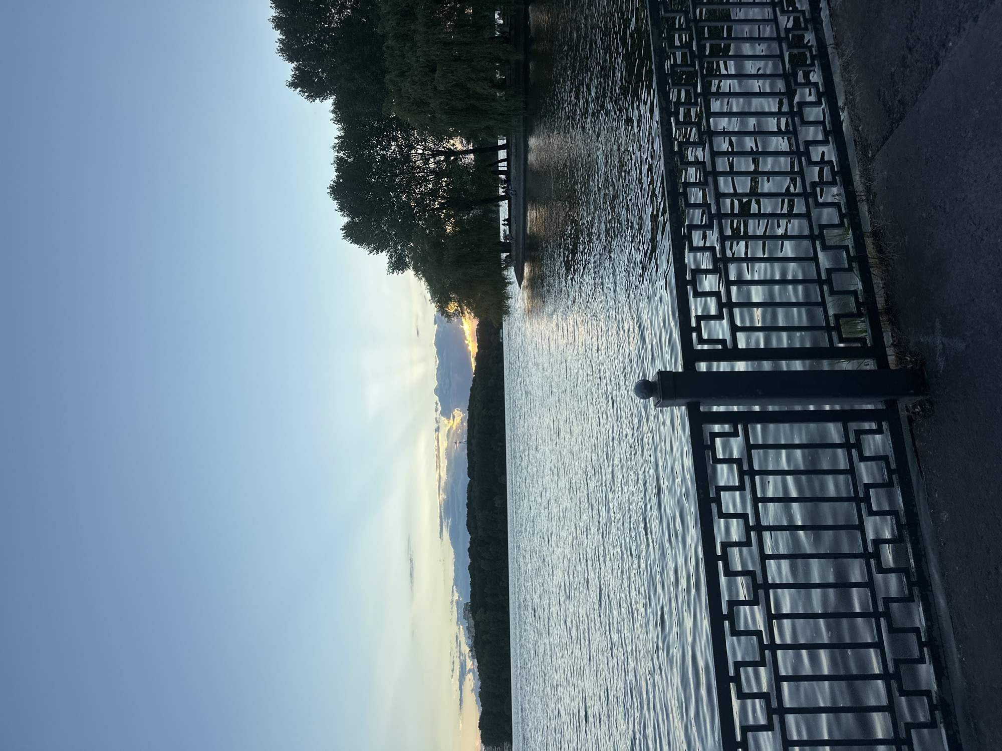 The image depicts a waterfront scene, likely Ternopil Lake in Ternopil, Ukraine, at dusk. A dark metal railing with a geometric pattern runs along the foreground, separating a paved walkway from the water. The lake's surface is rippled, reflecting the muted colors of the sky. In the distance, across the lake, is a tree-lined shore with a cluster of buildings visible on a slight incline. The sky above is a gradient of soft blues and grays, with rays of sunlight breaking through clouds near the horizon. A small group of people are seated or standing under trees on a grassy area by the water's edge in the mid-ground. The overall atmosphere is peaceful and serene.