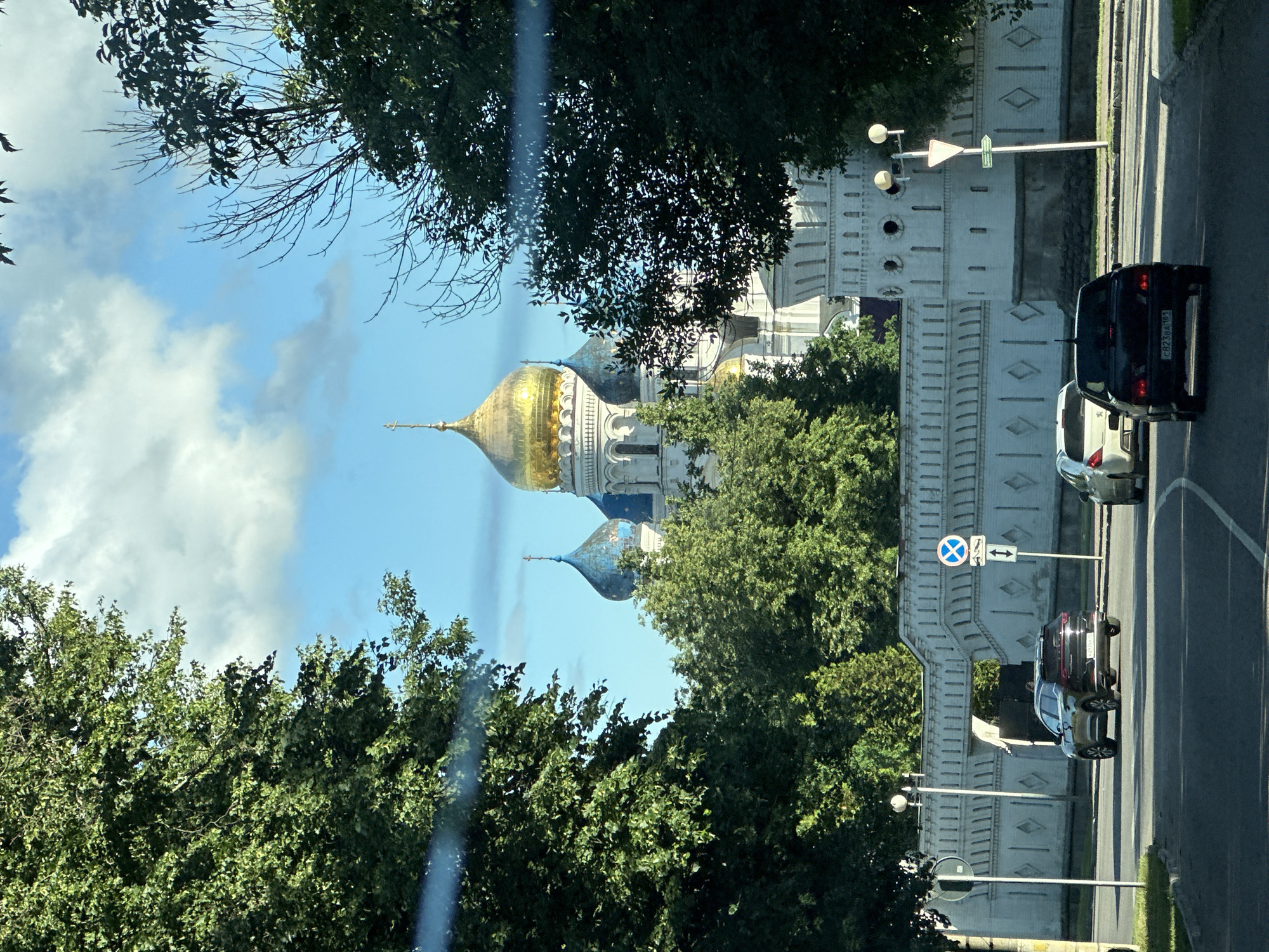 A sunny day in Dzerzhinsky, Russia, is depicted, with a view of the St. Nicholas Monastery. In the foreground, a black sedan and a white hatchback drive on a road. The black sedan is in the right lane, and the white hatchback is in the left lane, slightly ahead of it. The road is paved with asphalt and has white lane markings.
Behind the cars, a white stone wall with a patterned design runs along the street. Beyond the wall, the golden domes and blue spires of the St. Nicholas Monastery are visible, surrounded by lush green trees. The sky is blue with scattered white clouds. A traffic sign with a blue circle and a white cross is posted on the right side of the road, near the wall. Another traffic sign showing a white triangle with a red border is also visible on the right. The overall scene is one of a peaceful, sunny day in a city with historical and religious landmarks.