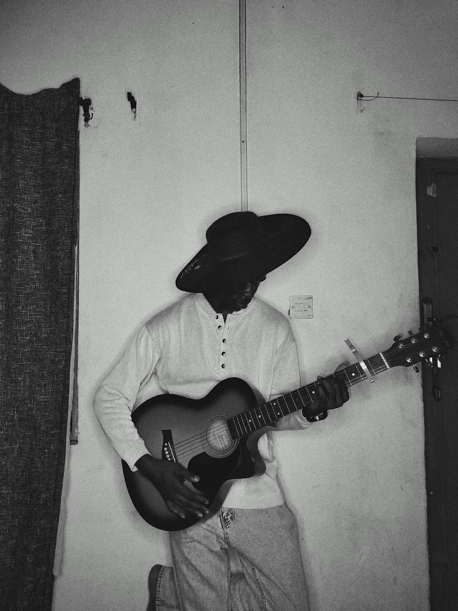 A person wearing a large-brimmed hat and a long-sleeved shirt is playing an acoustic guitar. The person is standing against a wall. To the left of the person, a curtain is visible. A light switch and door frame are seen on the right. The location is in the city of Unknown, Unknown.