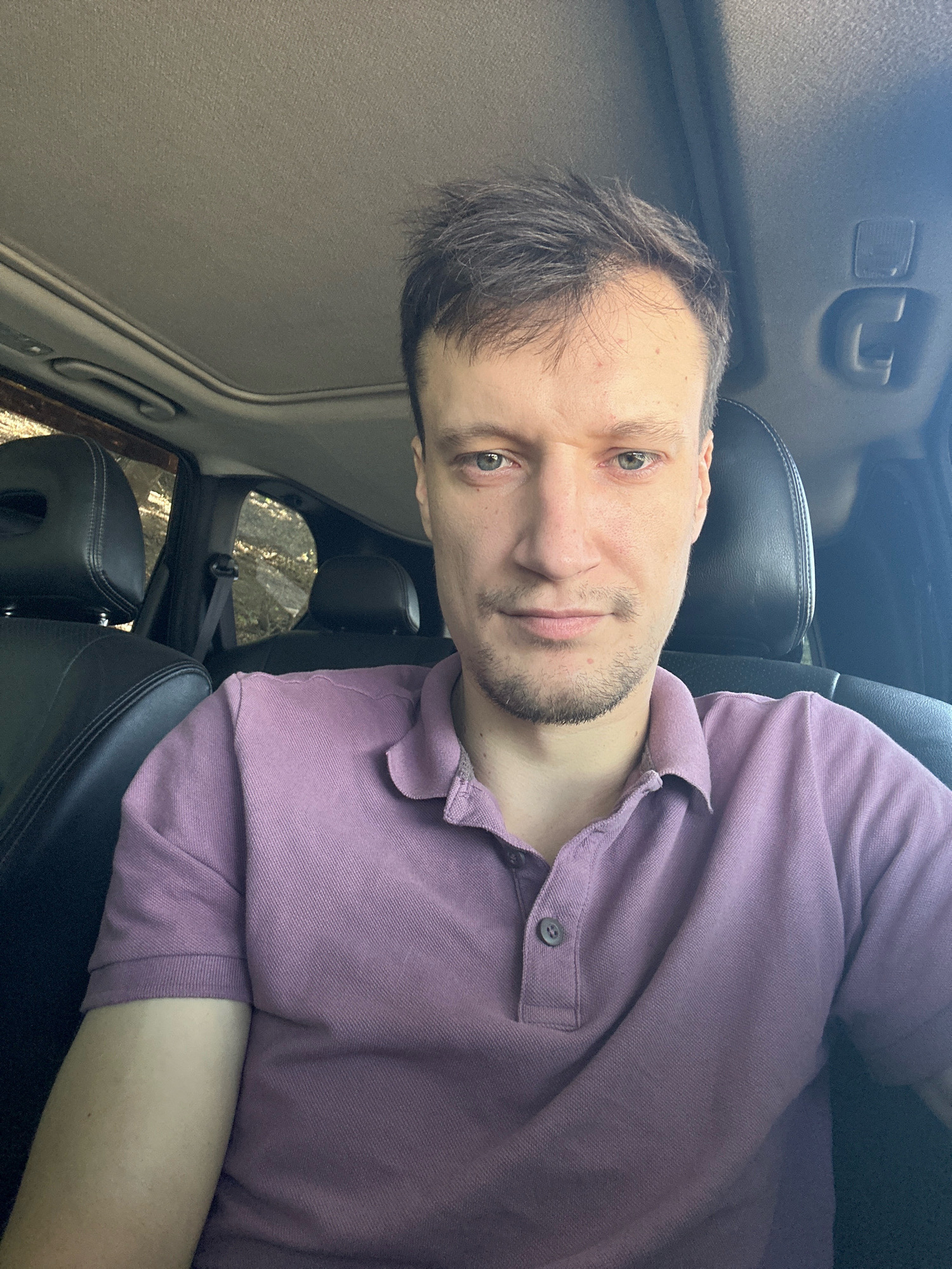 A man is seated in the driver's seat of a car, facing the camera. He has short brown hair, light-colored eyes, and a light beard. He is wearing a purple polo shirt. The car interior is visible, including the sunroof, leather seats, and seat belts. The background outside the car windows shows trees and foliage, suggesting an outdoor, possibly rural or suburban setting.
