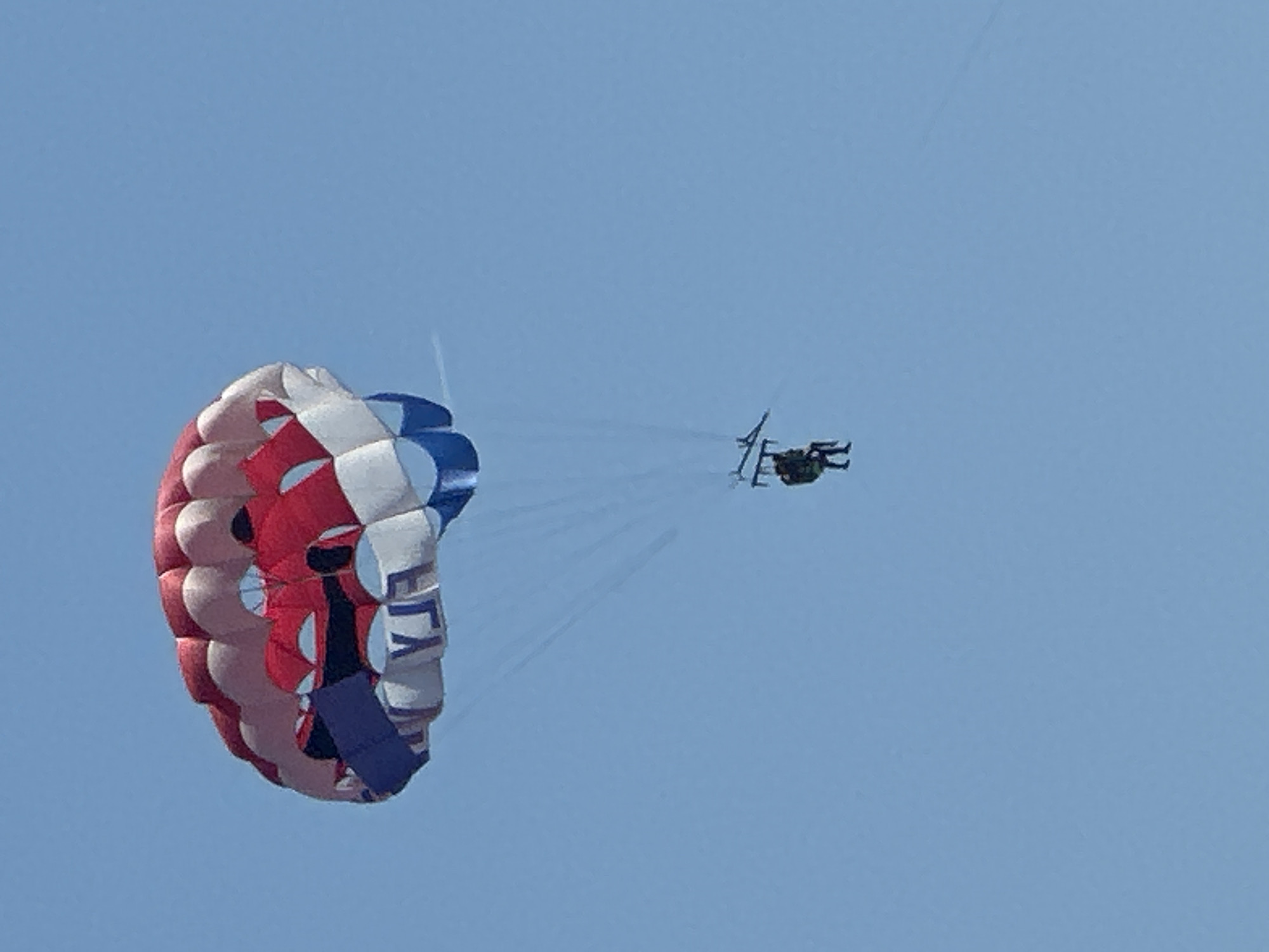 In a clear blue sky, a parasail with red, white, and blue sections is descending. The word "FLY" is visible on the underside of the parasail. Two individuals are suspended beneath the parasail, attached by harnesses and ropes. They are positioned centrally below the parasail. The scene is an outdoor activity. There is no visible indication of the city of Boreti, Montenegro, in the image.