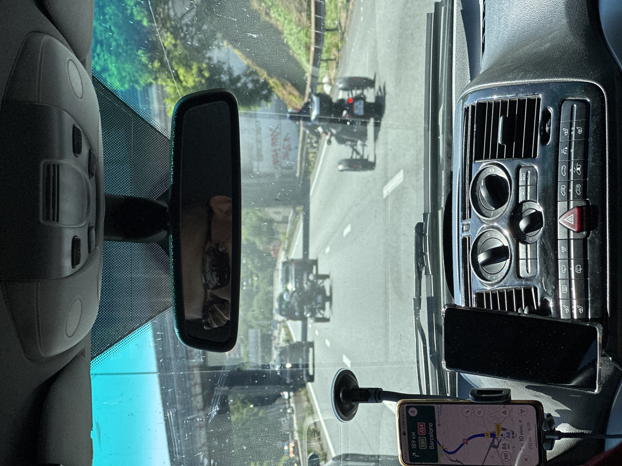 A view from inside a vehicle, possibly a car or van, looking out through the windshield. Several open-wheel race cars are visible on a road ahead. The interior of the vehicle is in focus, with the dashboard, rearview mirror, and phone mount with a visible GPS navigation app being clear. The app shows a route with the destination listed as "Barcelone" (Barcelona), with a distance of "189 км" (189 km) and an estimated time of arrival. There are two cell phones in view. One is running the GPS and another is resting on the dash. The photo was taken at an angle. The location is not in Villeneuve-Loubet, France. The GPS is headed to Barcelona.