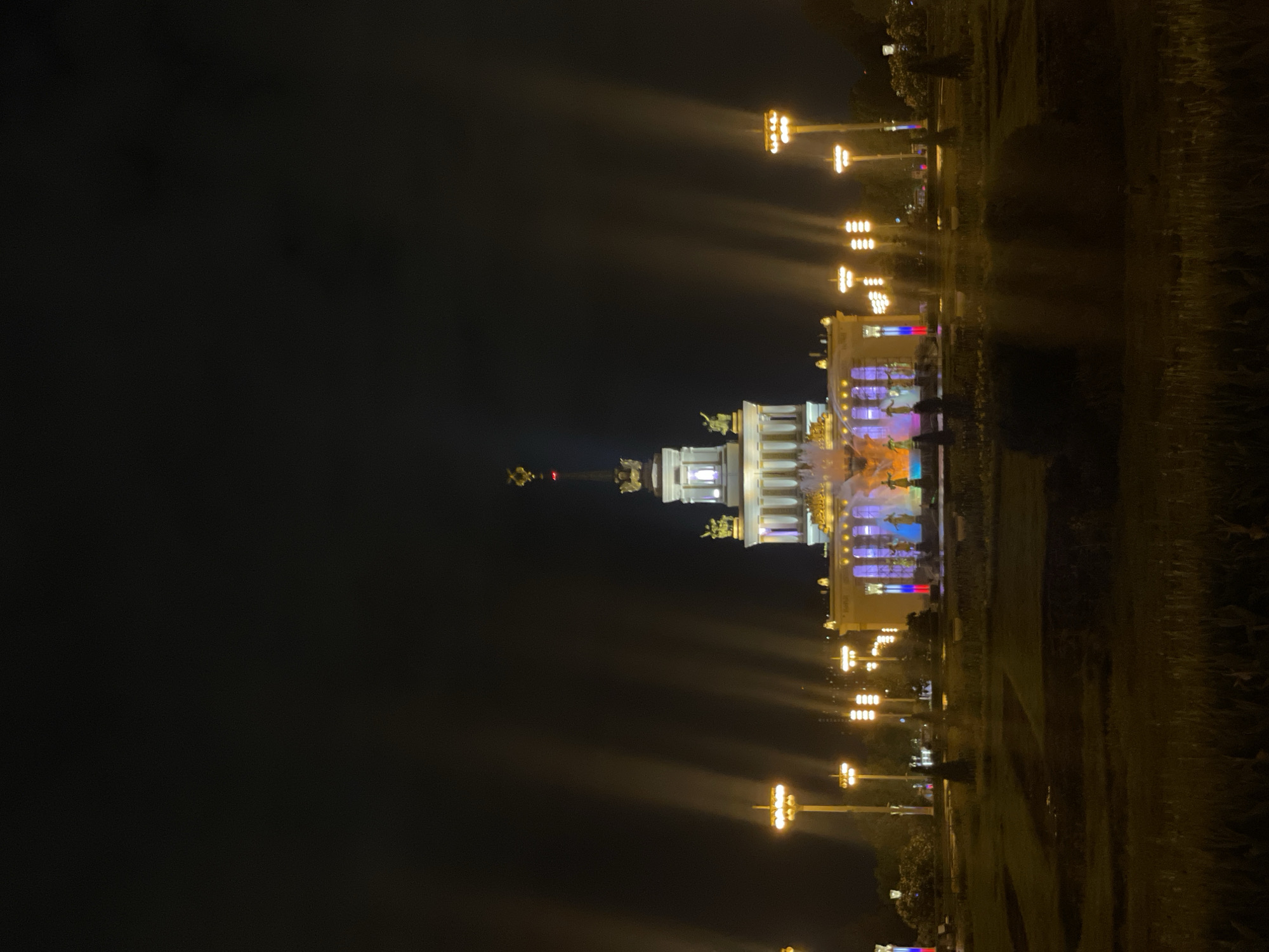At night, the Exhibition of Achievements of National Economy (VDNKh) in Moscow, Russia, is illuminated. A grand, neoclassical structure with multiple columns and sculptures, including the iconic Friendship of Nations Fountain, stands prominently. The fountain shoots water high into the air, lit up with colored lights. Street lights cast beams into the night, illuminating the manicured lawns and pathways surrounding the buildings and fountain. The scene is bathed in the glow of artificial light against the dark night sky.