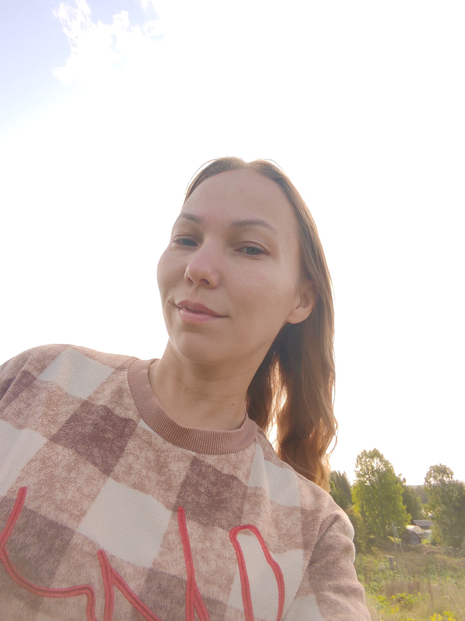 A young woman with long, light brown hair is captured in a close-up, outdoor selfie. She is looking towards the camera with a slight smile. She is wearing a brown and white checkered sweatshirt with a red embroidered design across the chest. The background is bright and slightly overexposed, suggesting a sunny day with a pale blue sky and scattered clouds. To the right of the woman, a rural landscape is visible, featuring green trees and some small buildings that suggest a village setting. The image appears to have been taken in Чухлэм, Russia.