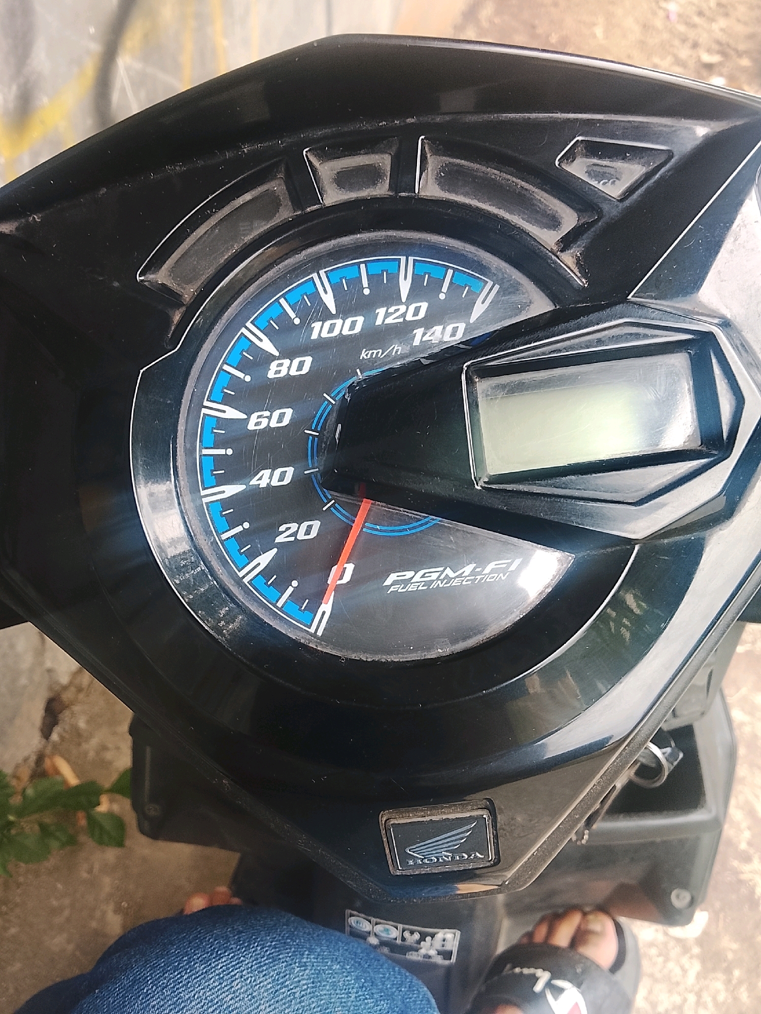 This is a close-up, top-down shot of a motorcycle's instrument cluster, featuring a speedometer and a small digital display. The speedometer has markings for 20, 40, 60, 80, 100, 120, and 140 km/h, with a red needle pointing between 0 and 20, indicating a speed below 20 km/h. Below the speedometer, the text "PGM-FI FUEL INJECTION" is visible. The digital display above the speedometer is illuminated and appears to be showing information, but the details are unclear. The instrument cluster is mounted on a black plastic surface. In the lower left and right of the image, parts of a person's legs are visible, wearing blue jeans and black sandals. The background consists of a textured surface, possibly concrete or asphalt, and some green foliage on the left. The image appears to be taken outdoors, likely in Kertahayu, Indonesia, given the context provided.