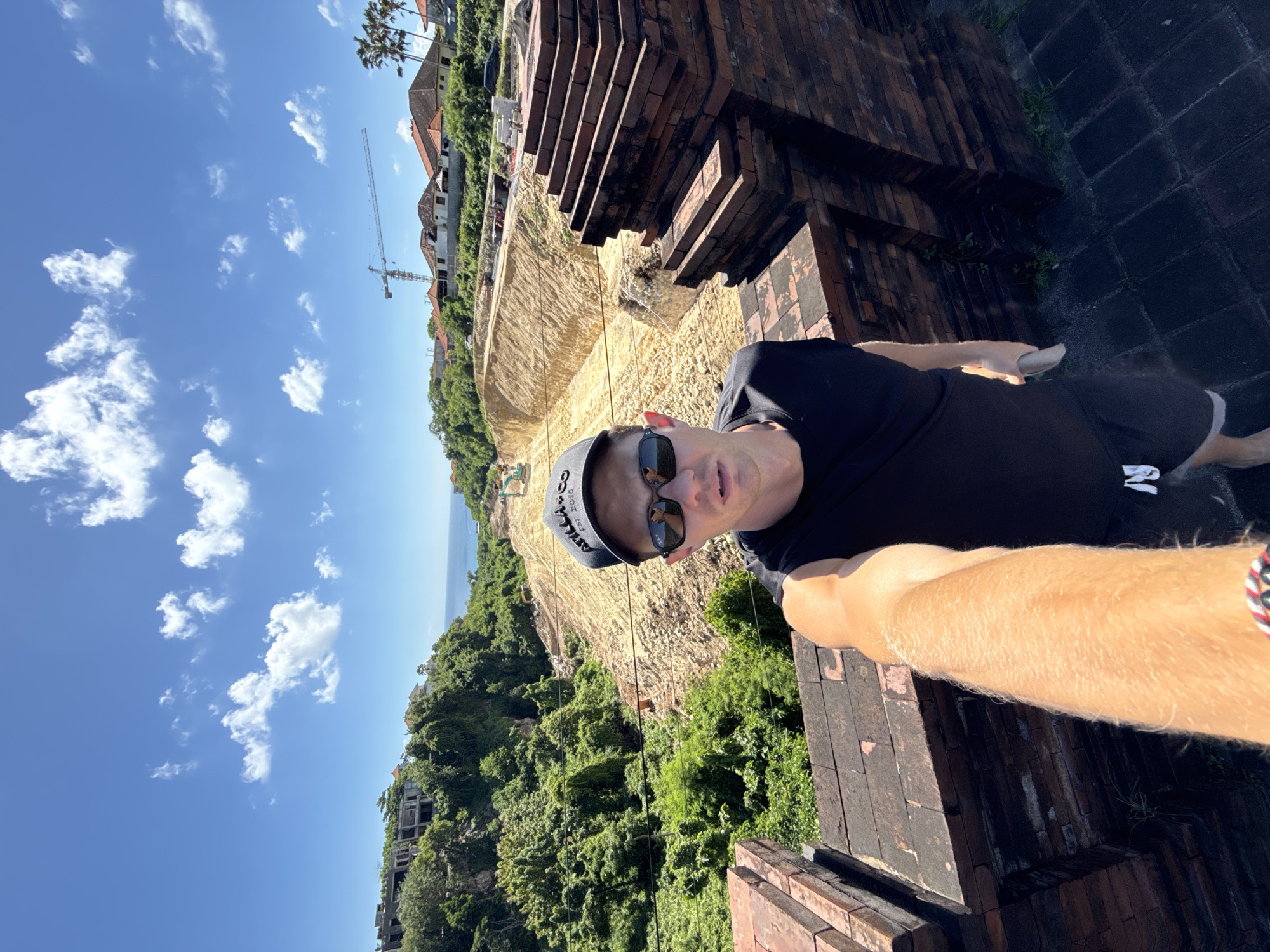 A male individual is captured in a selfie-style photograph, standing on what appears to be an elevated brick structure or wall. He is wearing a black t-shirt, dark shorts, sunglasses, and a dark baseball cap with a white logo and text "ST." (partially visible). His right arm is extended, holding the camera, and a patterned bracelet is visible on his right wrist. He looks directly at the camera with a neutral expression.
The setting is an outdoor, sunny environment. In the midground, behind the person, there's a large excavated area of bare, light brown earth, possibly indicating a construction site on a hillside or cliff. Several heavy machinery items, including what appears to be an excavator and a large construction crane, are visible in the distance within this area. Lush green tropical vegetation and trees densely cover the land, particularly lower down the slopes.
In the background, the vast blue expanse of the ocean is visible under a clear, bright blue sky dotted with scattered white clouds. Several residential buildings with red-tiled roofs are nestled among the greenery on higher ground, and palm trees are also present. The brick structure the person is standing on has a weathered appearance, suggesting an older construction. The visible text on the shorts includes the letter "R". The scene suggests a location undergoing development, possibly in a coastal, tropical region like Kuta Selatan, Indonesia.