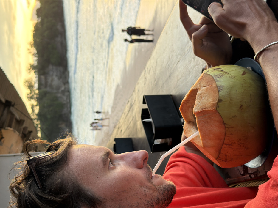 A male individual is seated at a dark wooden table in a beachside establishment, likely in Jimbaran, Indonesia, during sunset. He is depicted in profile, facing left, wearing a red short-sleeved shirt. Sunglasses are perched on his short brown hair, and he has light facial stubble. He is actively drinking from a large, partially peeled green-yellow coconut, which contains a white straw and rests on a blue plate. His right hand is holding and interacting with a black smartphone, while a thin silver bracelet adorns his right wrist. Two black containers are visible on the table behind the coconut. Beyond the table, a sandy beach extends to the ocean, illuminated by golden sunset light. Blurred figures are distributed along the shoreline; a group of approximately five individuals walk near the water's edge to the left, and two silhouetted figures stand further right. In the distant background, a tree-covered cliff face rises from the water.