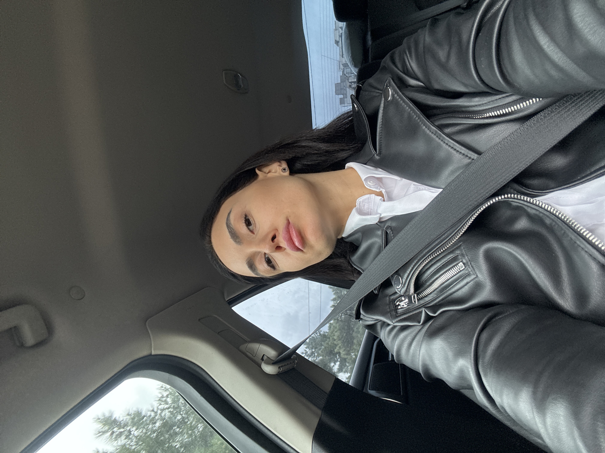 A person with dark hair is in the driver's seat of a car, wearing a black leather jacket over a white collared shirt and a seatbelt. The car interior is visible, including the window with a view of trees and buildings outside. The photo appears to be taken inside the car, with the camera angled slightly downwards. The specific location in Tbilisi, Georgia is not discernible from the image.