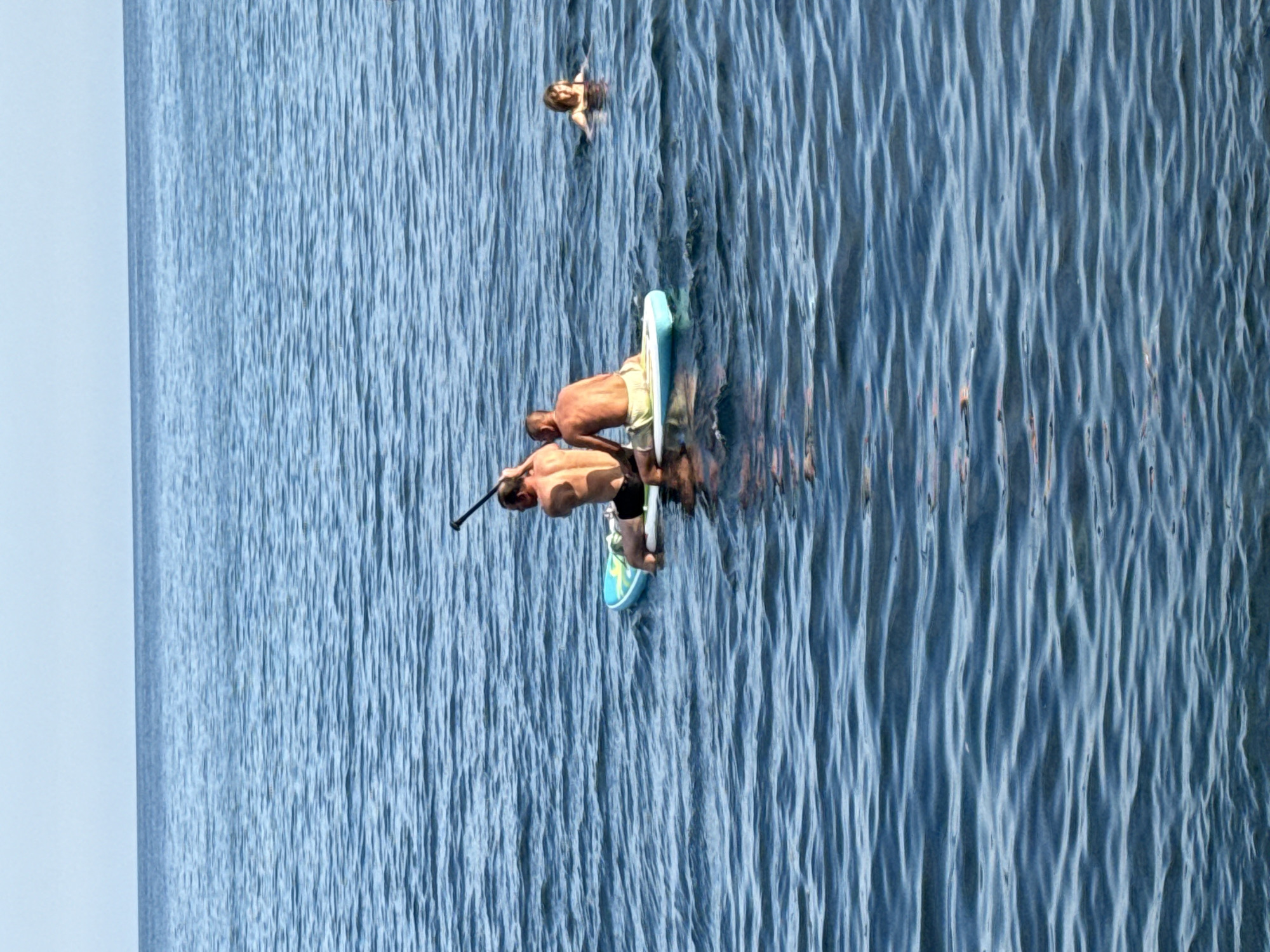 Two men are sitting on a paddleboard in the water. The man on the left is holding a paddle. The man on the right is wearing yellow shorts. A woman is swimming in the background to the right. The water is blue and choppy. The sky is clear and blue. This scene is in Utios, Ukraine.