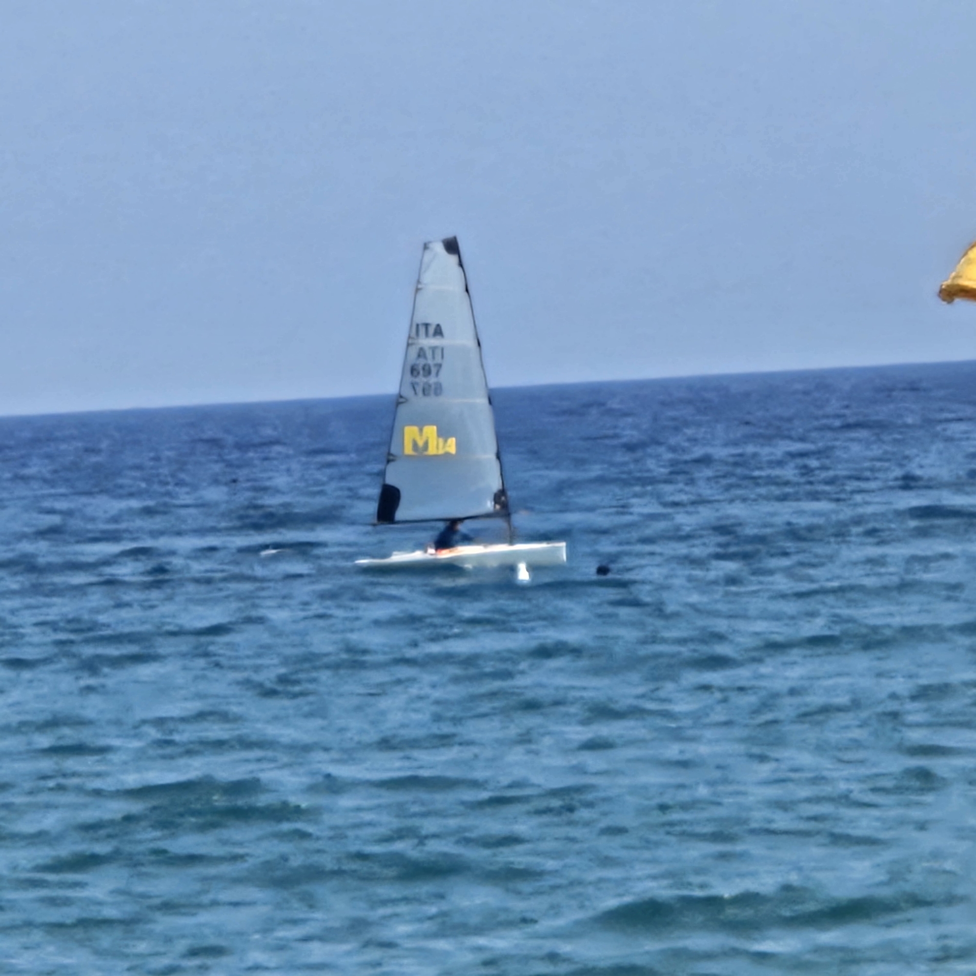 A sailing dinghy, identified by "ITA ATI 697 T00" and a yellow "M4" on its sail, is sailing on choppy blue water under a clear sky. A person, wearing a blue top, is visible in the dinghy. The horizon line is clearly defined. A small yellow object is visible in the upper right corner of the image. The scene is likely the Mediterranean Sea off the coast of Savona, Italy, given the "ITA" marking on the sail, which commonly denotes an Italian boat registration.