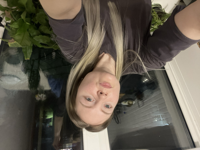 A selfie of a woman with blonde hair wearing a grey t-shirt. Her face is pale with blue eyes. Behind her is a window reflecting an outdoor scene at night. To the left of the woman are various green plants in pots. The window frame and a white wall are visible to the right. The photo is taken at an angle, causing the image to be rotated.