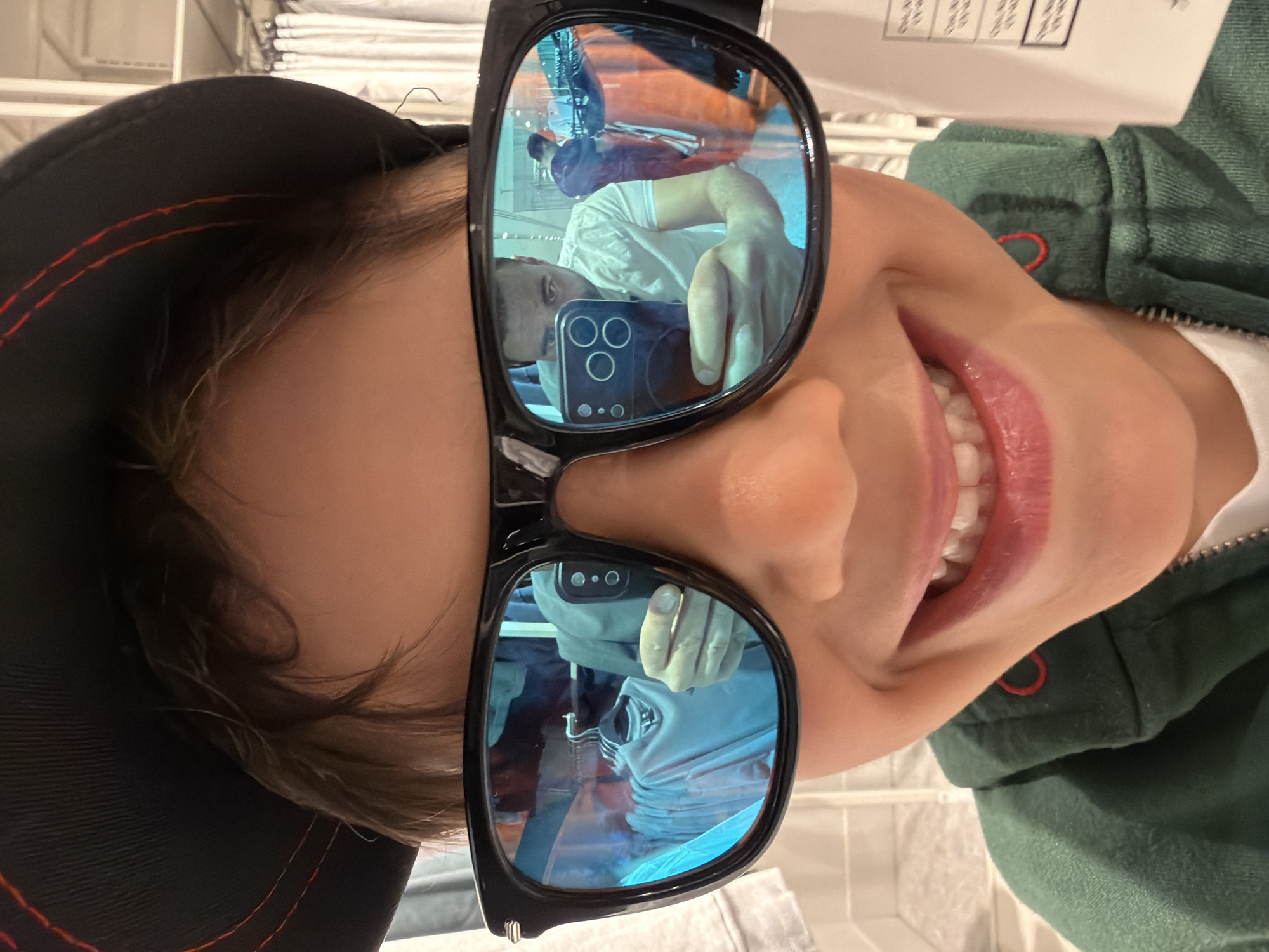 A portrait of a person wearing sunglasses and a hat, oriented upside down. The sunglasses reflect another individual holding a smartphone taking the picture. The person is wearing a green jacket, possibly a hoodie. The picture appears to have been taken in a retail setting, with folded goods or merchandise visible in the background. The reflection in the glasses suggests the photographer is in a similar setting. It is not possible to determine the location in London from the image.
