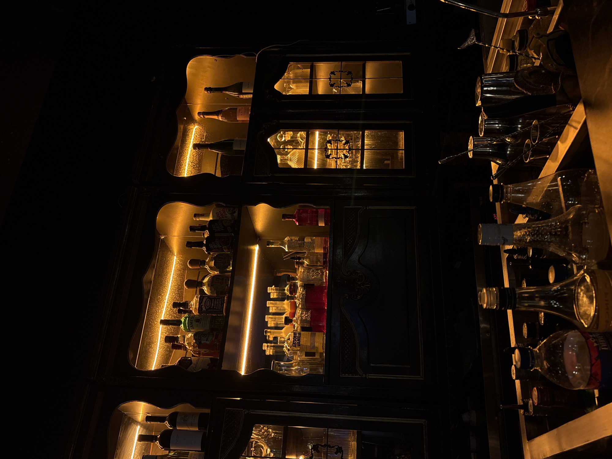 This image depicts the interior of a dimly lit bar, located in Saint Petersburg, Russia. A large, dark wooden bar cabinet with ornate carvings dominates the scene, featuring multiple shelves illuminated by warm yellow LED strip lighting. The upper shelves are stocked with numerous liquor bottles, including identifiable brands such as Absolut Vanilia, Jameson, Jack Daniel's, Chivas, Ballantine's, Whitley Neill Gin, and some champagne or wine bottles, including Calvet. Glass-fronted cabinets on the left and right sides of the main display contain various types of glassware, also subtly illuminated. The lower section of the image reveals the top of a bar counter. On the counter and within a recessed metallic well, numerous bottles, some fitted with pour spouts, are present. A collection of metallic bar tools, including shakers, measuring jiggers, strainers, and bar spoons, are arranged within the well. No individuals are visible in the scene.