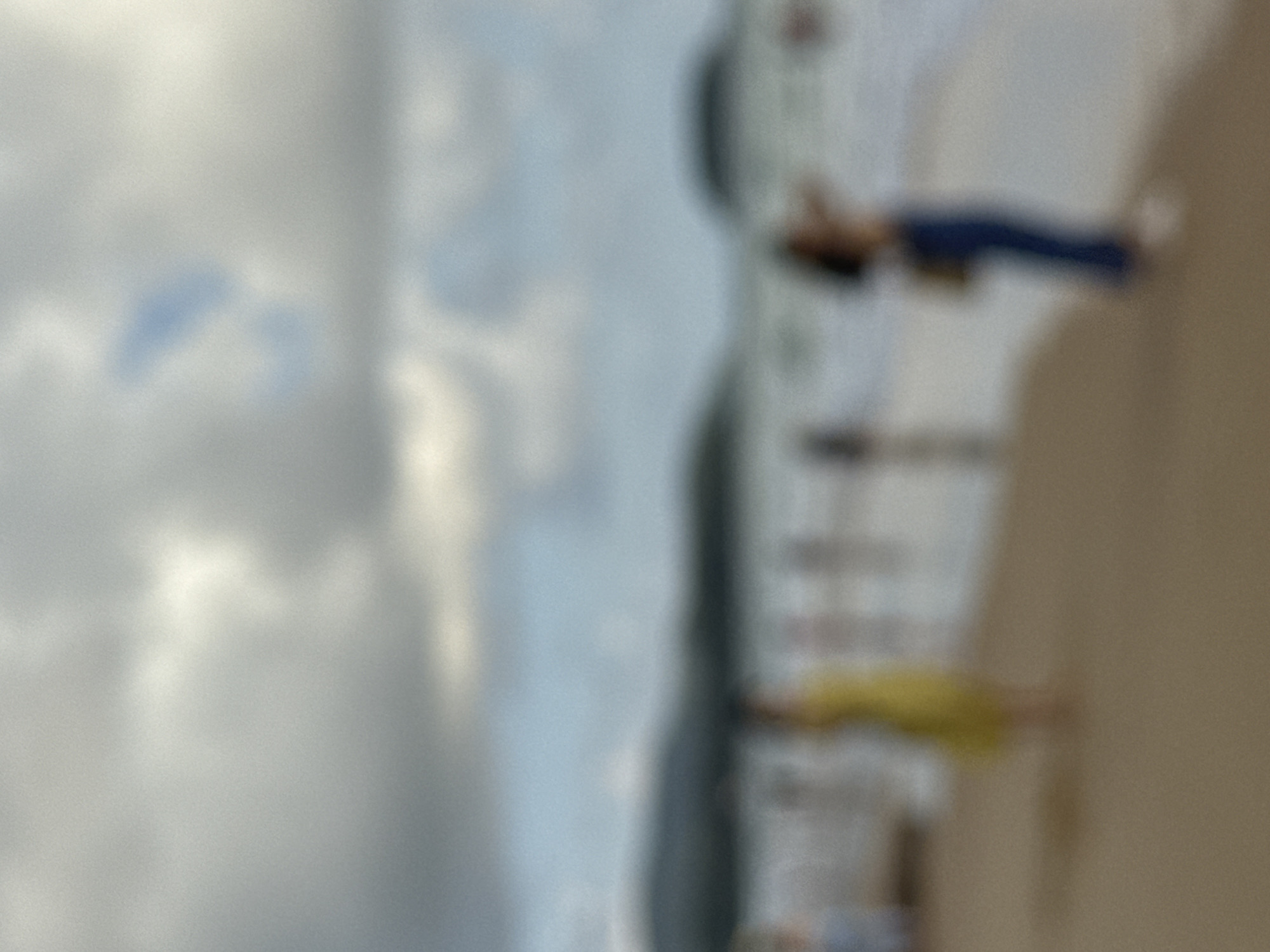 The image shows a blurred scene of a beach, likely in Karon, Thailand. In the foreground, two figures are visible on the sandy beach. The figure on the left is wearing a yellow dress. The figure on the right is wearing blue pants and a dark top. Both figures appear to be standing and facing towards the water. The middle ground shows the ocean with waves, and further in the background, there are blurry shapes that could be hills or landmasses. The sky is overcast with grey and white clouds, with some patches of blue sky visible. The overall impression is a somewhat hazy and indistinct beach environment.