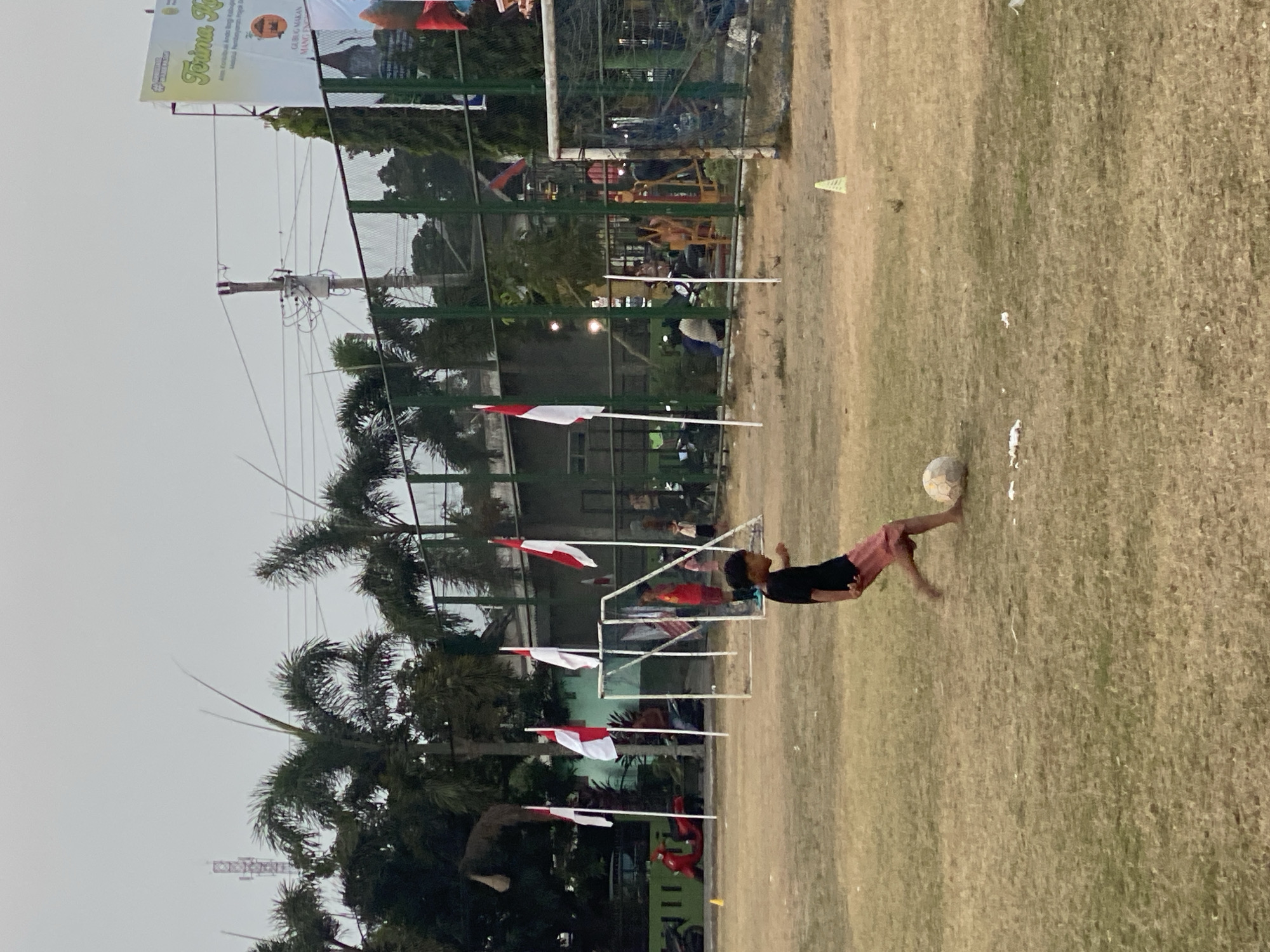 A boy in a black t-shirt and pink shorts plays soccer on a dry, grassy field. He is kicking a ball, captured in motion with his right foot. In the background, several Indonesian flags on poles are visible, suggesting a national celebration or event. A soccer goal is set up on the right side of the frame, and another incomplete goal structure is in the middle ground. Beyond the field, there are trees, a fence, and some buildings. A large banner with text is also visible in the upper right corner. A red scooter is parked on the left side of the image. The sky is overcast. This scene is likely taking place in Kasihan, Indonesia, given the context of Indonesian flags.
