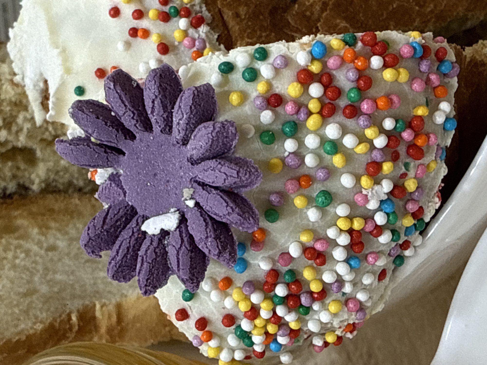 A close-up shot depicts a decorated cupcake with white frosting. Multi-colored sprinkles cover the frosting, and a large purple flower adorns the cupcake's side. Sections of other baked goods are visible in the background. The location is unknown and cannot be determined from the image. There are no people visible.