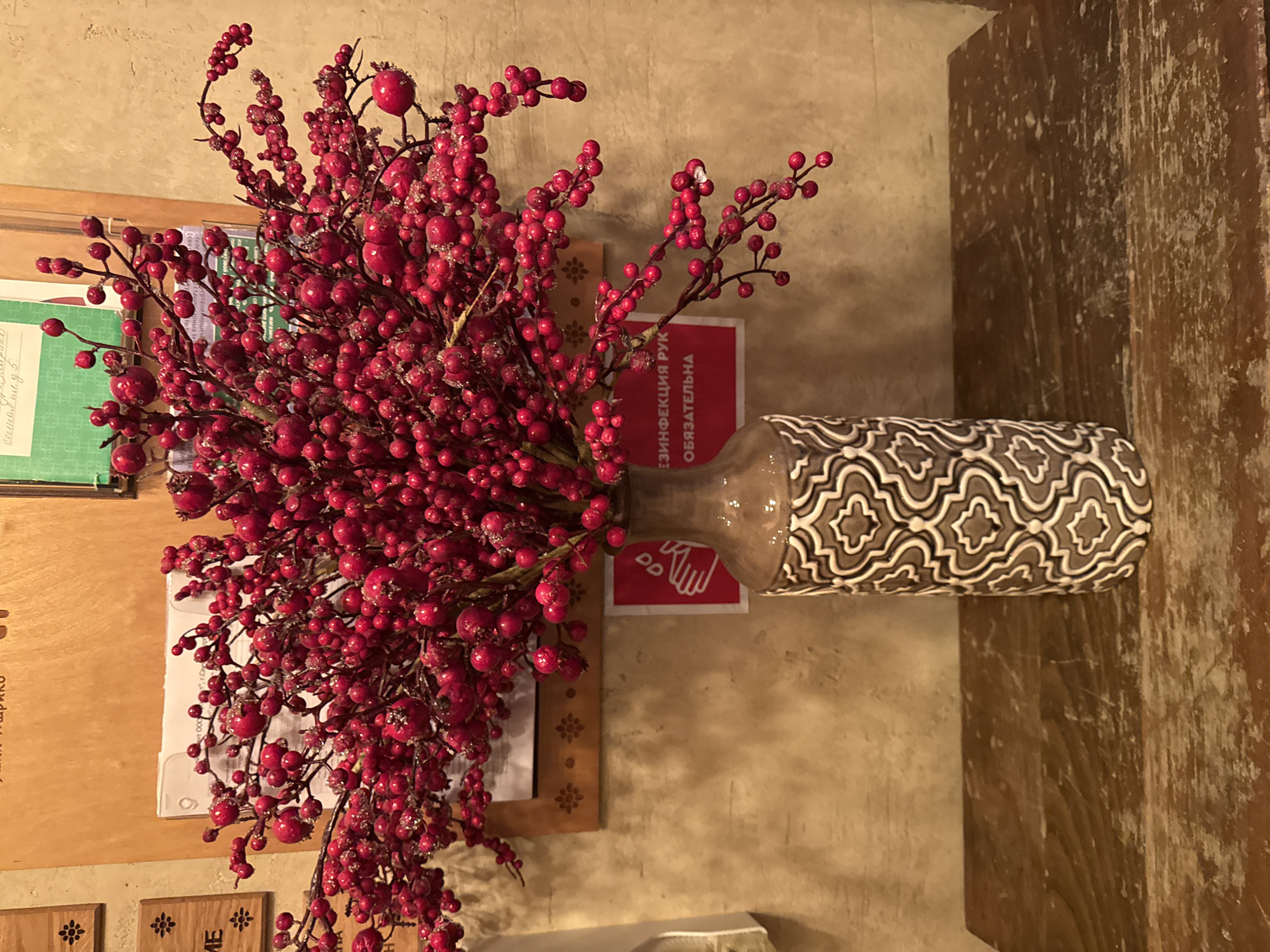 A bouquet of artificial red berries, some with white, crystalline embellishments, is displayed in a decorative, patterned vase on a distressed wooden surface. The vase is cylindrical with a narrow neck and features an intricate geometric pattern in shades of brown and white. Behind the vase, a textured, light brown wall is visible. To the left of the vase, a small wooden plaque with a carved flower motif is mounted on the wall. Further left, another wooden plaque with the Cyrillic letter "E" and a floral design is also present. A piece of paper with text and some green paper are affixed to the wall behind the berry bouquet. A red sign with a white hand icon and Cyrillic text, which translates to "Disinfection of hands is mandatory," is partially visible to the right of the vase. The overall lighting suggests an indoor setting. The location is identified as Saint Petersburg, Russia.