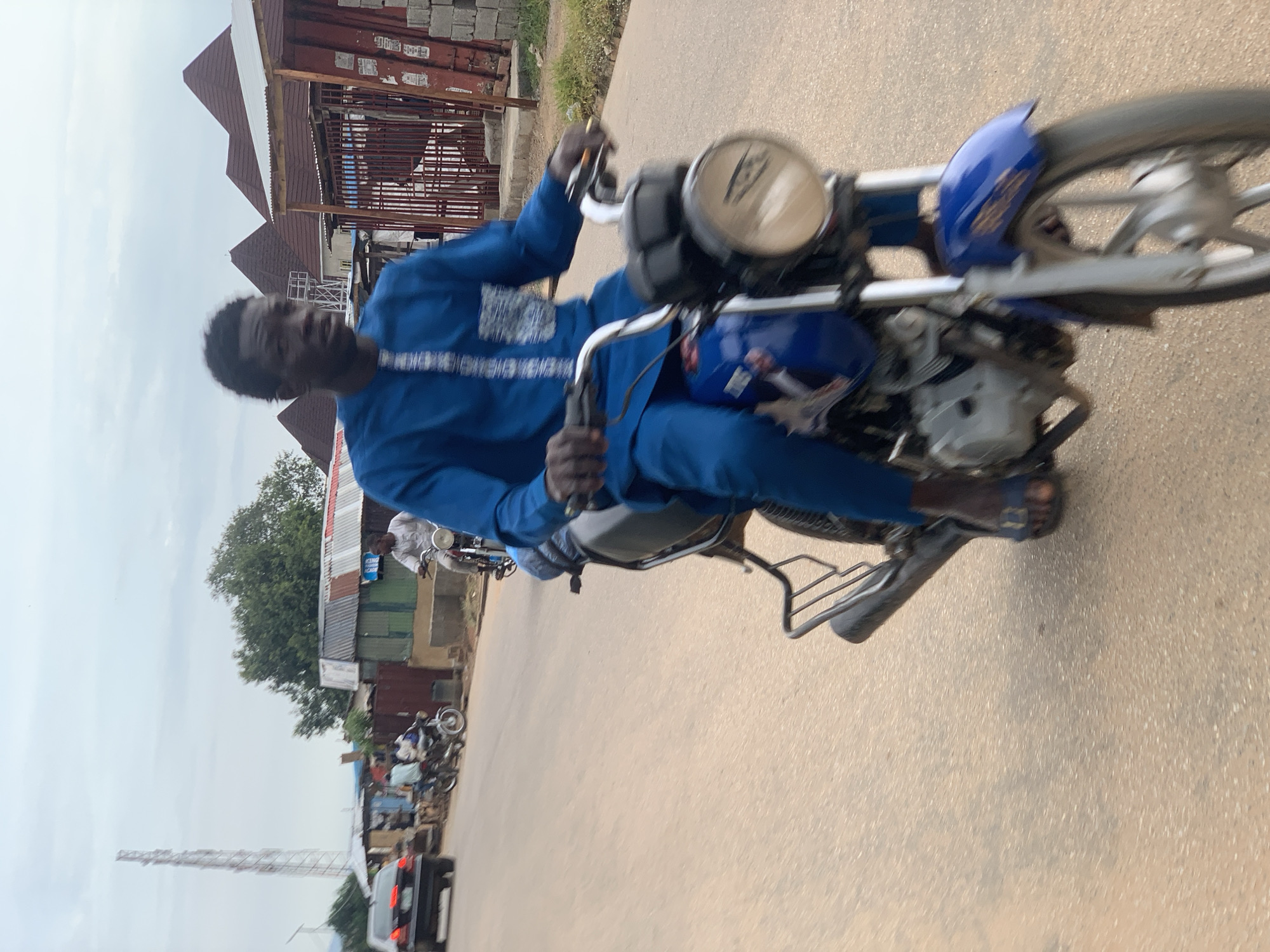 A person is riding a blue and black motorcycle on a paved road in Abuja, Nigeria. The rider is wearing a bright blue, long-sleeved traditional outfit with a white, patterned embellishment on the chest. They are looking forward and their hands are on the handlebars. The motorcycle has a silver exhaust pipe, a black seat, and a silver engine. The road appears to be made of asphalt and is a light brown color. In the background, there are several buildings with tin roofs, some of which are painted in green and brown. There are also other vehicles on the road, including a black car and other motorcycles with riders. A tall telecommunications tower is visible in the distance. The sky is overcast with light grey clouds. The image has a slight motion blur, indicating the motorcycle is in motion.