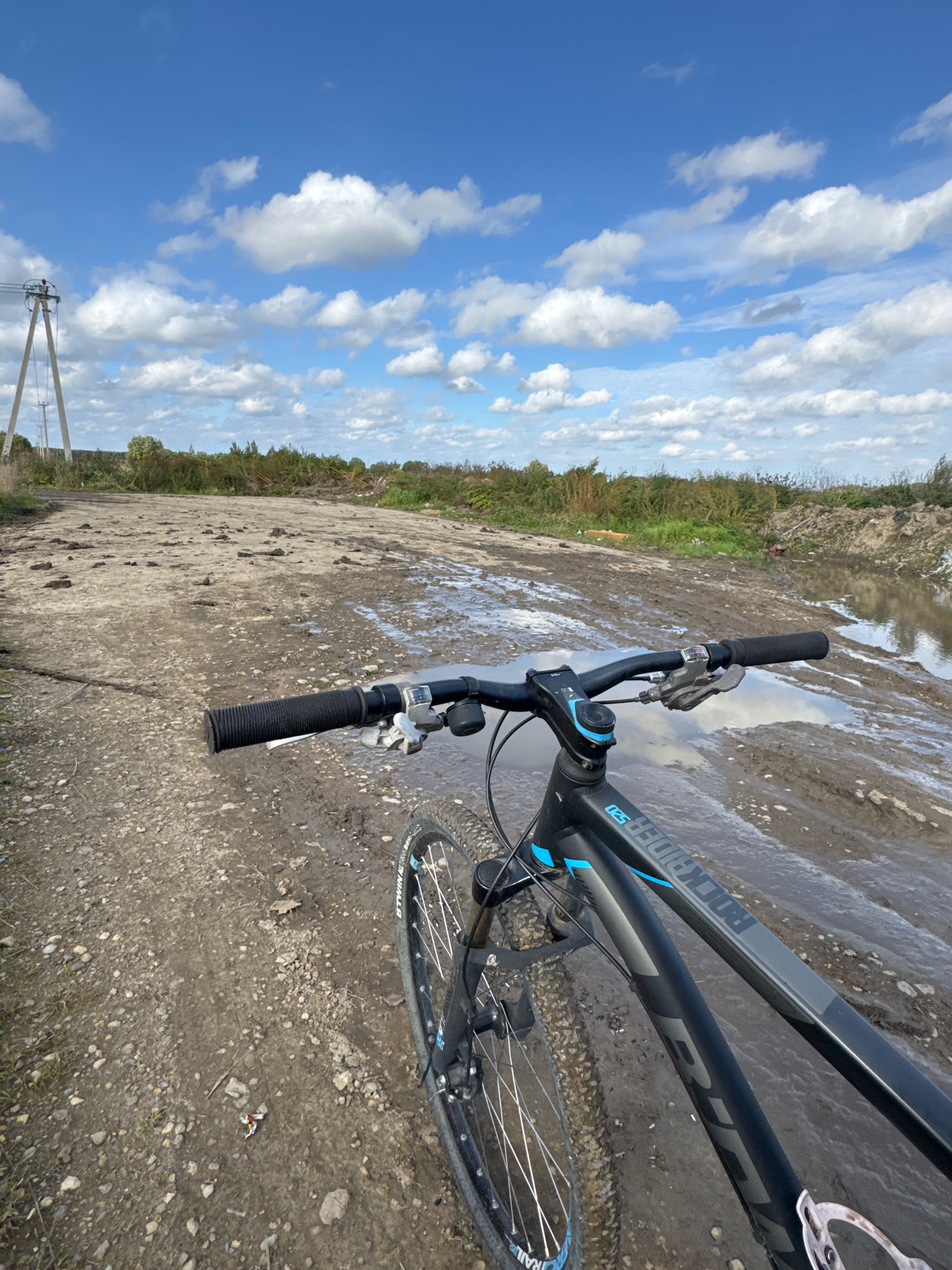 A first-person perspective shot from a bicycle shows a muddy, unpaved path under a partly cloudy sky. The bicycle's handlebars, stem, and front wheel are visible in the foreground. The path is wet and muddy, with several puddles. In the background, there is vegetation and a power pole on the left side of the image. The scene appears to be located in an undeveloped area, possibly on the outskirts of a rural or industrial zone. No people are visible.