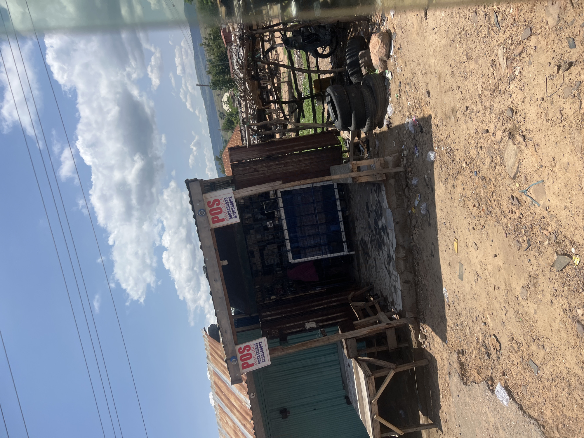 The image displays an outdoor scene in Chum, Nigeria, under a clear blue sky with scattered cumulus clouds and multiple overhead power lines. The foreground consists of unpaved, sandy-brown earth littered with small rocks, plastic fragments, and other debris. A patch of broken, lighter-colored concrete is visible on the far left foreground. Shadows cast by structures suggest bright daylight.
In the midground, a small, open-fronted commercial structure is visible, constructed with a rusty, corrugated metal roof and wooden supports. Two red and white signs are affixed to the front, each displaying "POS AVAILABLE" and phone numbers "5083433533" and "8250586092." The entrance area of this structure is paved with black and white patterned tiles. Inside, a person wearing a dark pink or maroon top is partially visible.
To the immediate left of this main structure stands another building with a matching rusty corrugated metal roof and a green corrugated metal wall. A raw wooden table or stand is positioned directly in front of this building.
To the right of the main commercial structure, a rustic enclosure constructed from wooden poles and branches is visible. Within this enclosure, a black motorcycle is parked. Outside the enclosure, on the dirt ground, several used tires are stacked and individually scattered.
In the background, distant hills or elevated terrain are visible, sparsely covered with vegetation and dotted with a few distant buildings, all beneath the expansive sky. A green tint along the right edge of the image suggests it was captured through a vehicle window.