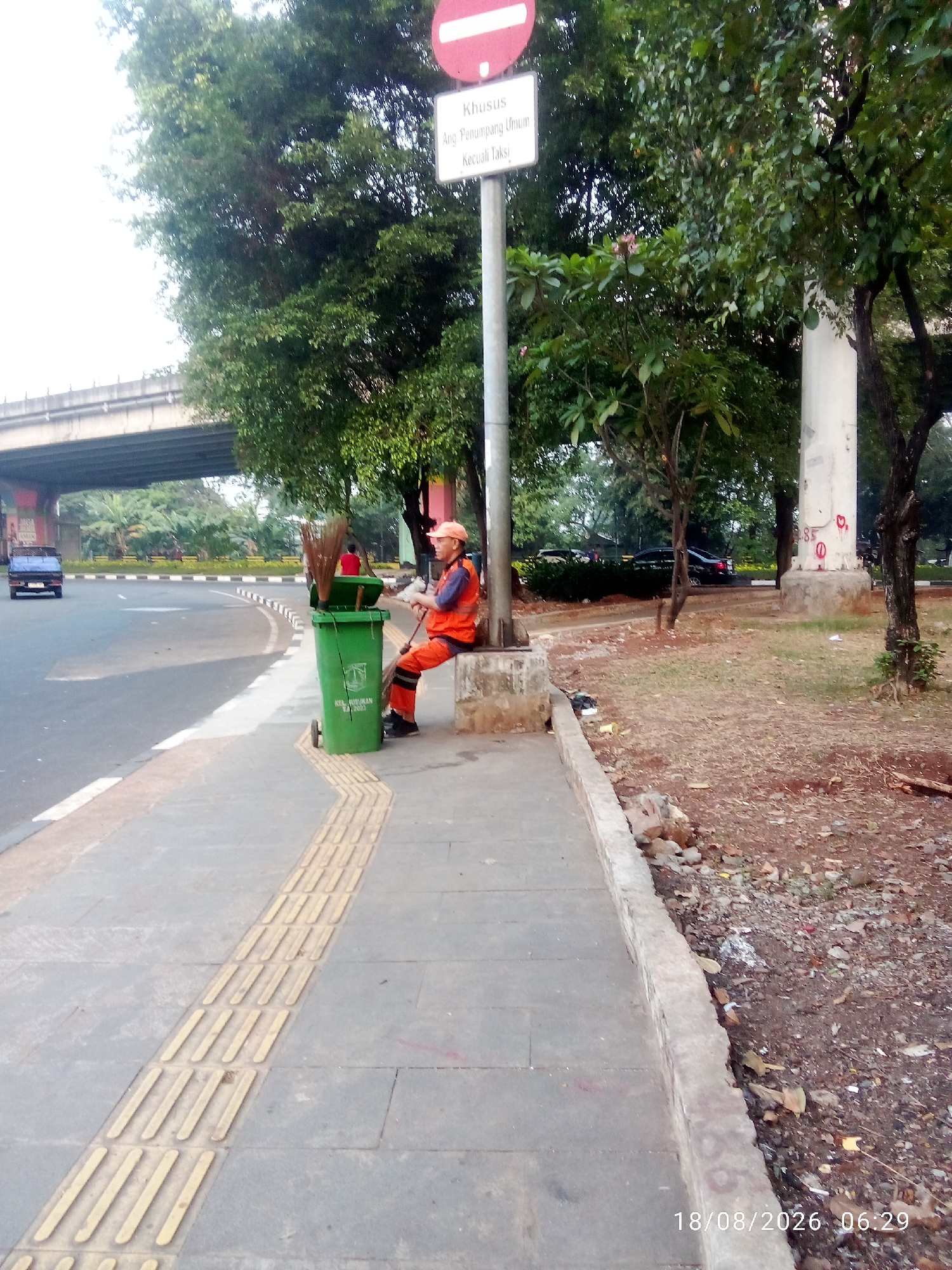 A street cleaner in an orange uniform and cap sits on a concrete bench next to a green trash bin on a sidewalk in East Jakarta, Indonesia. The cleaner is holding a broom and appears to be taking a break. A traffic sign with a red circle and a white bar is visible behind him, along with trees and a road with a blue pickup truck driving away. The sidewalk has a tactile paving strip for visually impaired pedestrians. The date and time stamp "18/08/2026 06:29" is visible in the lower right corner of the image.