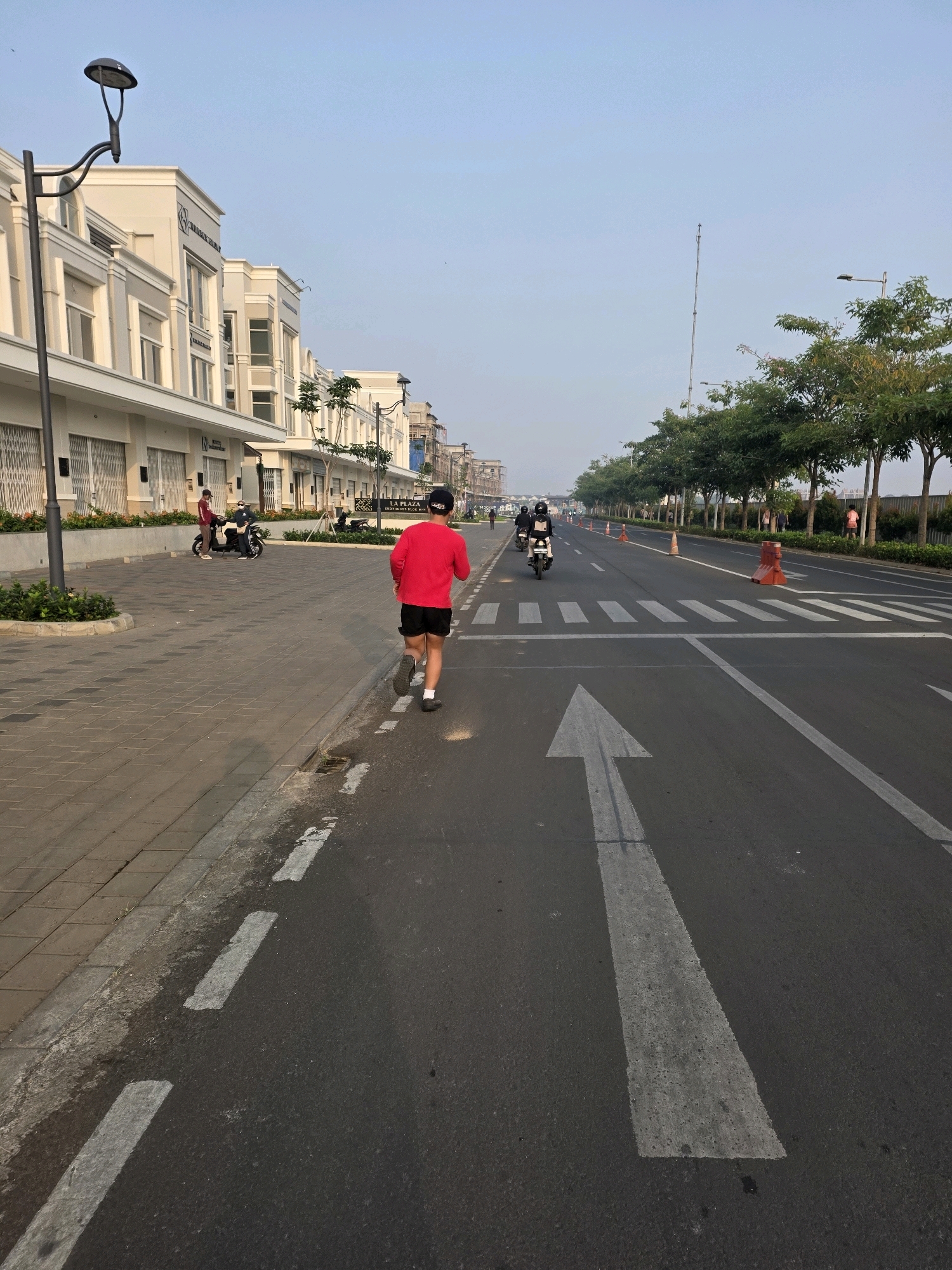 In Bojongsoang, Indonesia, a person wearing a red shirt and black shorts jogs along a paved sidewalk next to a road. The road is marked with white lane lines and a pedestrian crossing. Several motorcycles are visible on the road, some with riders. Buildings with modern architecture line one side of the street, while trees and vegetation are on the other. A lamppost stands near the sidewalk. The sky is clear and pale blue.