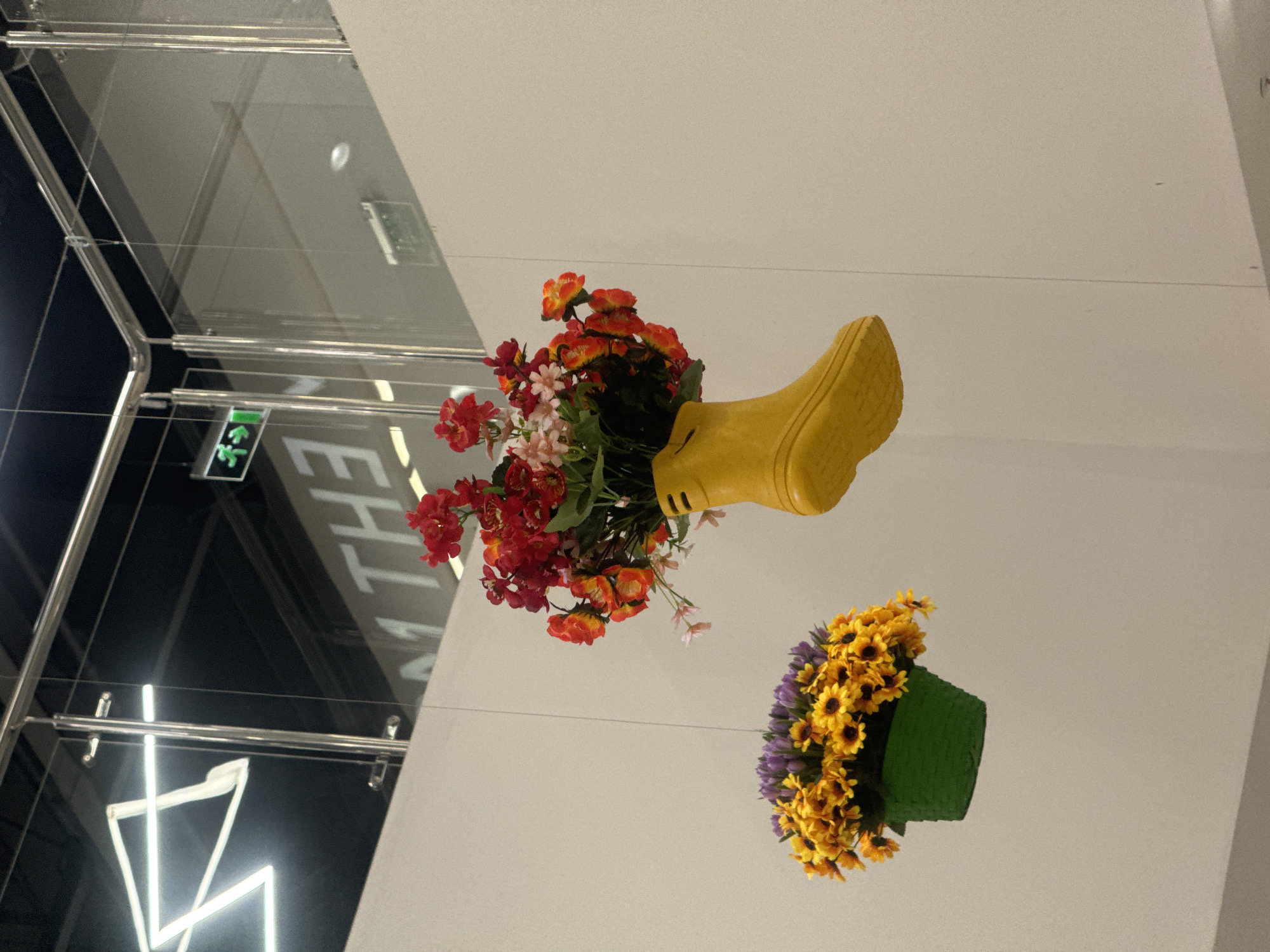 This image shows a bright yellow Wellington boot filled with red and orange flowers hanging from a wire. To the left, a green planter filled with yellow sunflowers and purple flowers also hangs from a wire. Both are suspended in what appears to be an indoor space with a white wall and a ceiling that has a metallic railing and glass panels. In the background, there is a large sign with white lettering that reads "M THE". There is also a green exit sign with a running figure. This image does not contain any elements that would suggest its location in Irkutsk, Russia.