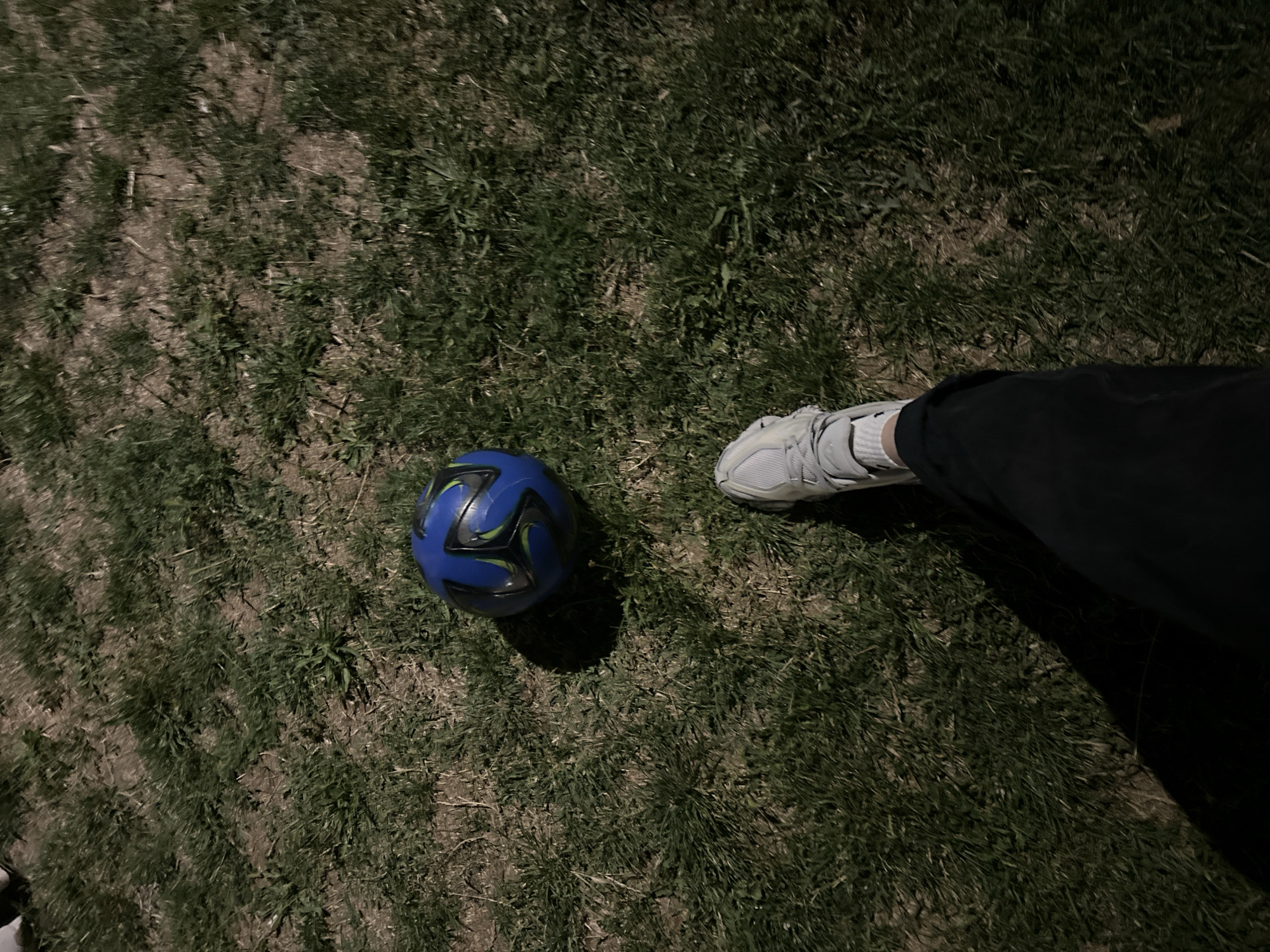 A person's legs and a soccer ball are visible on a patch of grass, viewed from a high angle. The person is wearing light grey sneakers, white socks, and dark pants. The soccer ball is blue with green and black accents. The scene appears to be outdoors, possibly in a park or yard, with uneven terrain and dry patches of grass. The lighting suggests it is either early morning or late evening. The location is identified as Unknown, Unknown.