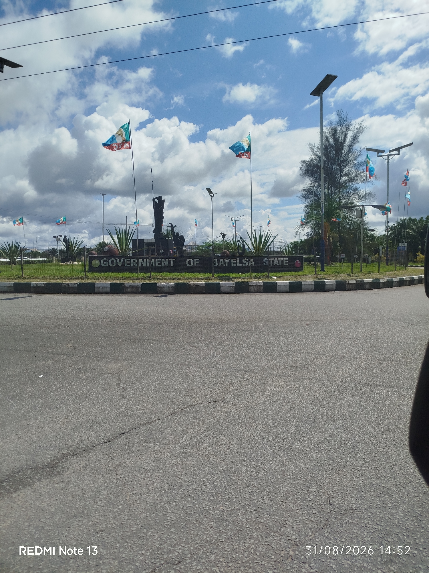 The image shows a roadside view of the "GOVERNMENT OF BAYELSA STATE" sign in Yenegoa, Nigeria. The sign is prominently displayed with large, black, block letters against a dark background, bordered by green grass and a striped curb. Behind the sign, a statue of figures stands tall, with several flags of varying designs flying on poles. The sky is partly cloudy, with a mix of white and grey clouds against a blue backdrop. Several streetlights, some equipped with solar panels, are visible. The foreground features a paved road with visible cracks. A timestamp at the bottom right reads "31/08/2026 14:52". A watermark "REDMI Note 13" is present at the bottom left.