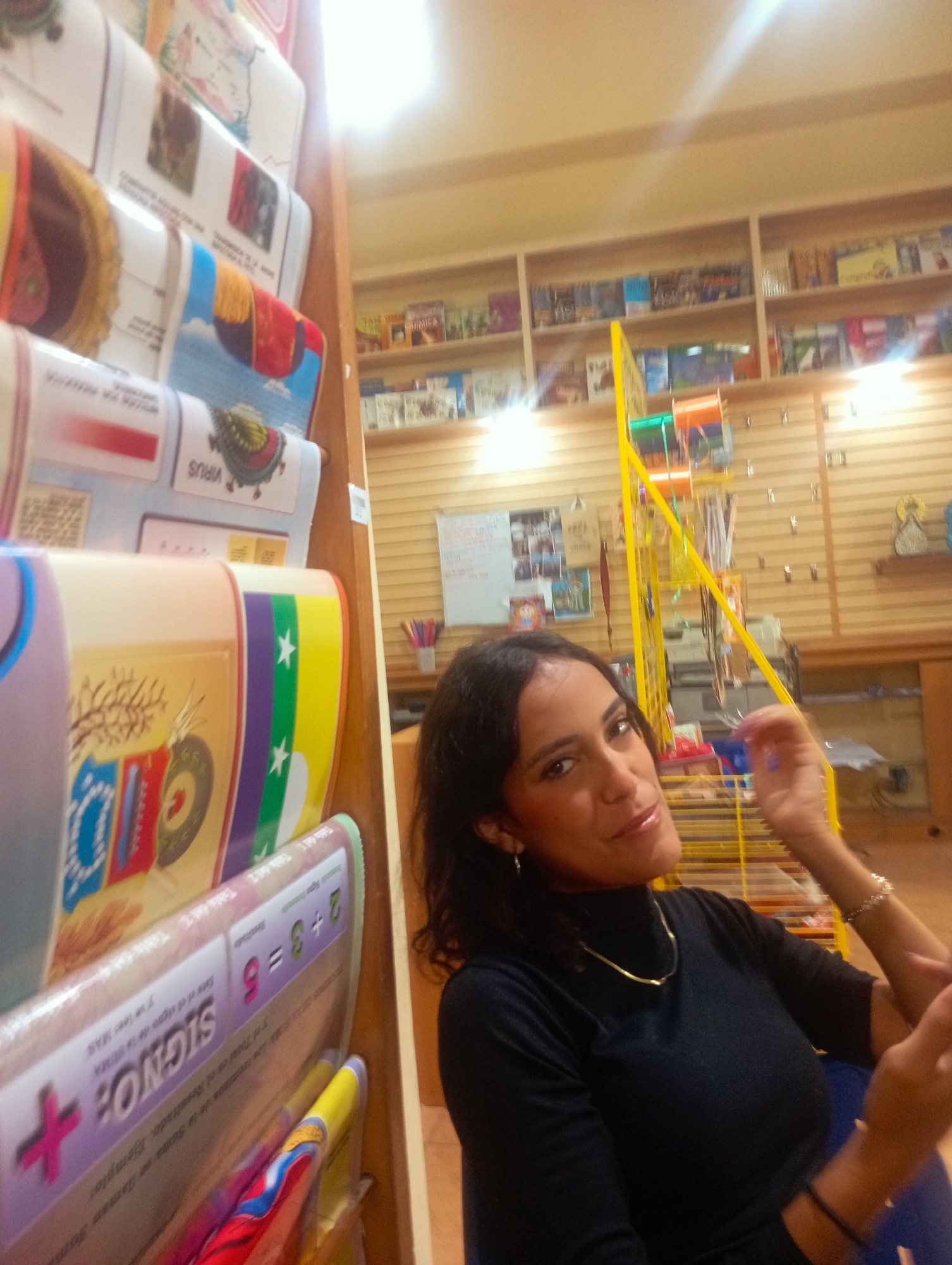 An eye-level, medium shot shows a woman with dark hair and makeup posing in what appears to be a bookstore or educational supply store. She is wearing a black turtleneck and a gold chain necklace. The woman is looking at the camera with a slight smile and her right hand is raised. In the background, shelves are filled with books and educational materials. A yellow shelving unit with colorful items is visible behind her. The overall lighting suggests an indoor setting.
