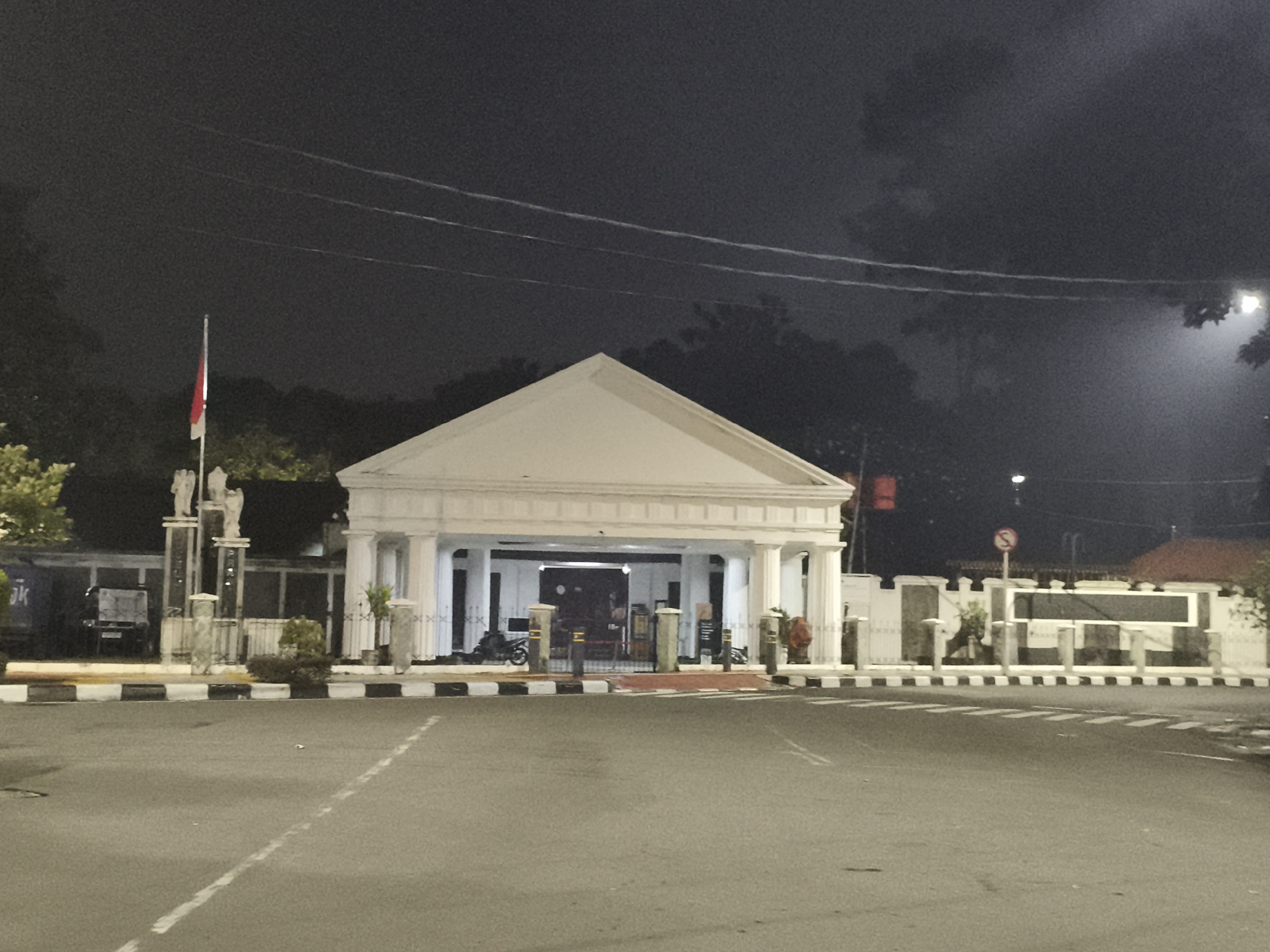 Captured at night, the image presents the exterior of a white building featuring a classical architectural design with columns and a triangular pediment. The building is positioned behind a zebra-striped barrier and an expanse of pavement. Inside the building's open portico, a motorcycle and a red barrier are visible. To the left of the building stands a flag pole displaying the Indonesian flag. Adjacent to the flagpole are two sculptural columns. Street lighting illuminates the scene, though the overall ambiance is dim, and the sky is dark and overcast. A "No Entry" sign can be seen in front of the building. The location is likely in Central Jakarta, Indonesia.