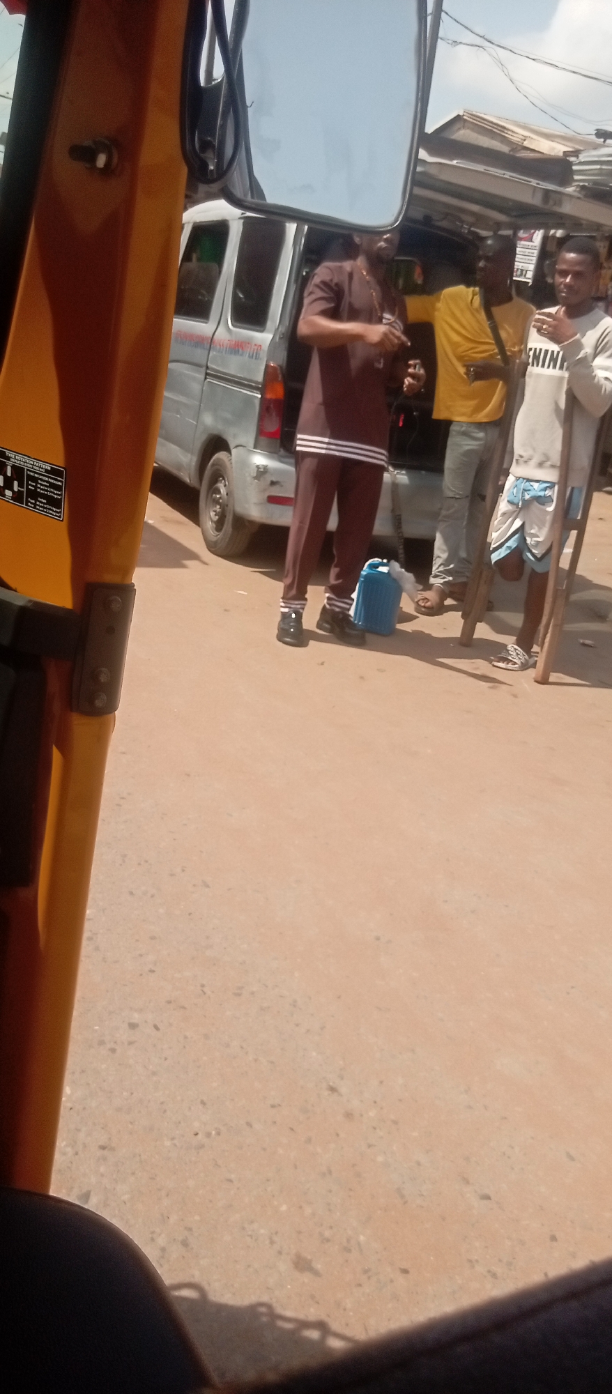 The image shows three men standing near a silver van in what appears to be a street scene. One man is wearing a brown two-piece outfit with white stripes on the pants and a gold chain. Next to him is a man wearing a yellow shirt and gray pants. The third man is wearing a gray shirt with "BENIN" printed on it and patterned shorts, and he is using crutches. A blue jerrycan sits on the ground near the man in brown. The van is parked on a paved street with a building or structure visible in the background. Part of an orange vehicle, possibly a tricycle taxi (keke), is visible in the foreground. The location is presumed to be Onitsha, Nigeria.