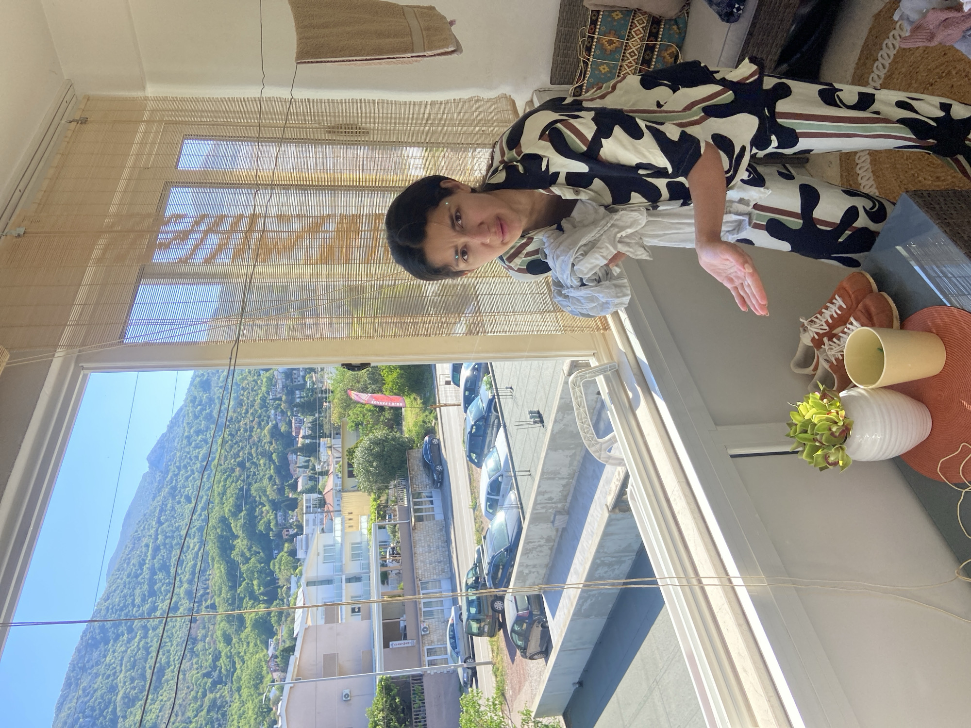 A brightly lit indoor scene looks out onto a sunny, mountainous landscape, featuring a woman standing near a window. The image, originally rotated, is best viewed with the woman on the right and the exterior view on the left.
The woman, with dark hair pulled back, wears a distinctive jumpsuit or two-piece outfit adorned with large, organic black and off-white shapes, accented by a thin reddish-brown stripe. Her expression is neutral as she looks towards the camera. She holds a crumpled white cloth in her left hand, which rests on a surface, while her right arm extends slightly with an open palm. Small, light-colored dots are visible above and between her eyebrows.
The room features a large window with slatted wooden blinds in the upper section, casting striped shadows on the white wall. To her right, various items are arranged on a surface: a pair of brown sneakers, a cream-colored ceramic mug, a white ribbed vase holding artificial green plants, and an orange woven mat. Further back, part of a patterned rug and a woven light-colored rug are visible on the floor, along with a brown towel draped over furniture.
Outside the window, a lush, green, tree-covered mountain dominates the background under a clear blue sky. In the mid-ground, numerous white and light-colored residential buildings dot the landscape. A road with several parked cars is visible, as well as a concrete structure, possibly a parking area, with more vehicles. Utility lines crisscross the exterior view. A red flag outside displays partial, reversed text, likely "HOTEL". The shoes have "ONITSUKA TIGER" faintly visible on the side. The scene depicts a sunny day in a coastal mountainous town, consistent with Sutomore, Montenegro.