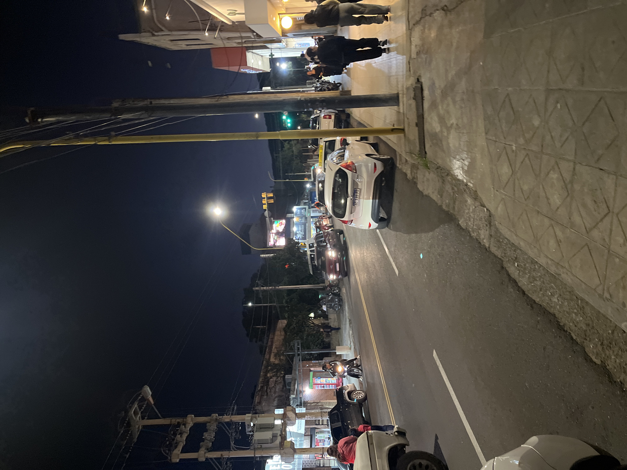 The image depicts a bustling night scene on a street in San Fernando del Valle de Catamarca, Argentina. The sky is dark, indicating nighttime, with artificial lights from streetlamps, vehicle headlights, and storefronts illuminating the area.
On the right side of the frame, a concrete sidewalk runs alongside a paved road marked with a solid white line and a double yellow line. A white car, possibly a hatchback, is parked on the street, displaying the Argentine license plate "AG 158 PH." Further down the road, several other vehicles, including cars and motorcycles/scooters, are visible, some parked and some in motion. A person is seen riding a scooter further down the street. Pedestrians are present on the sidewalk, with two individuals walking away from the camera in the upper right.
To the left, large utility poles with a dense network of electrical wires crisscross the frame. A prominent, bright street lamp casts light onto the middle-left area. Buildings with illuminated storefronts line both sides of the street, suggesting commercial activity. A traffic light in the distance shows a green signal, while another closer traffic light displays yellow. In the bottom-left, there's a utility pole with complex electrical infrastructure.
Notable details include the clear nighttime setting and the active urban environment. A textured concrete wall with a distinctive diamond pattern is visible along the right edge, separating the sidewalk from what appears to be a lower section. Visible text includes the license plate "AG 158 PH" and partially obscured text on a distant storefront sign that appears to read "PRESIDEN...", "TELEFONO PA...", and possibly "24 HS" below it, hinting at a business service. The scene captures the typical evening activity of a city street.