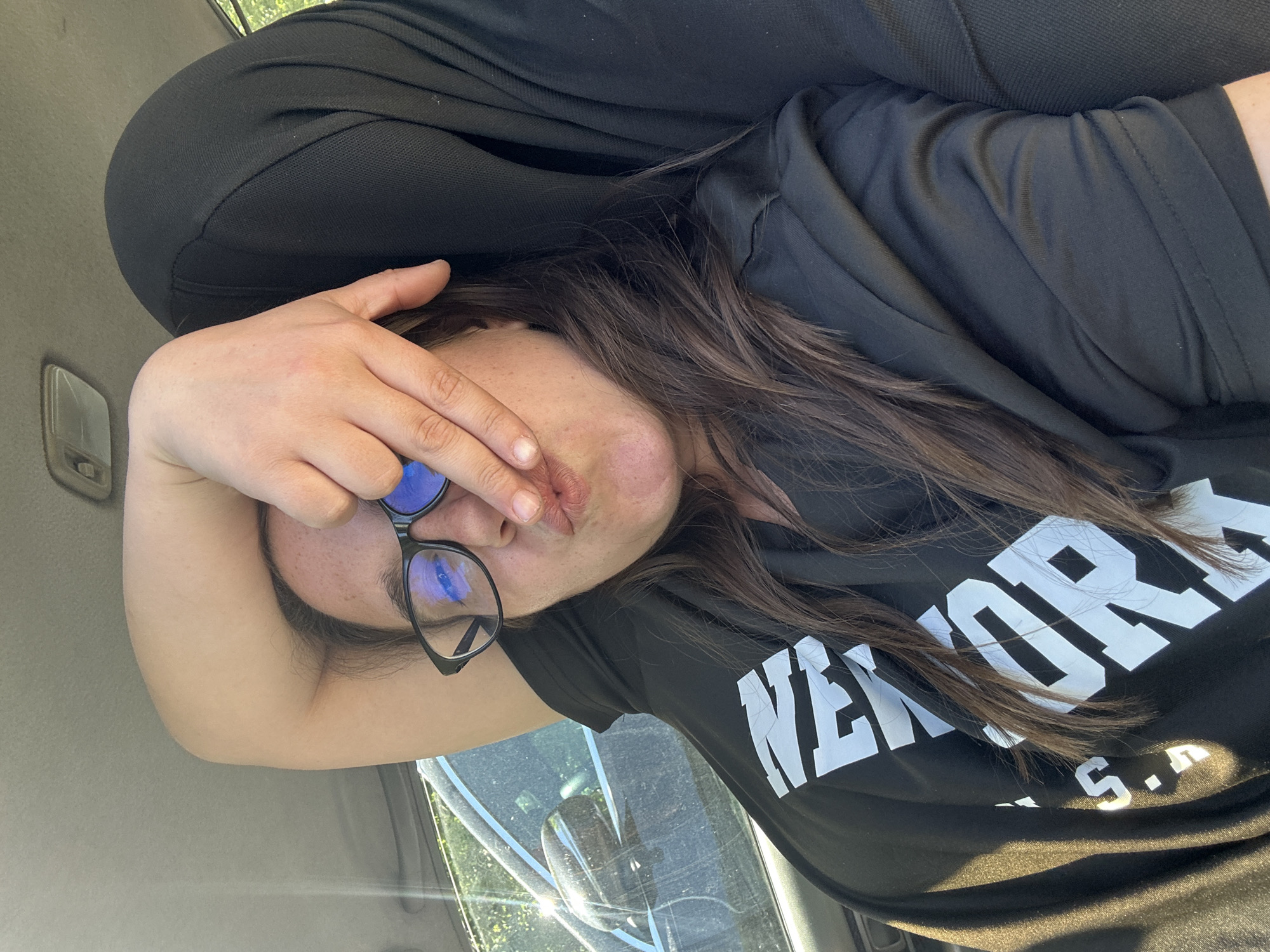 A woman is seated in a car, shielding her face with her hand. She has medium-length brown hair and is wearing glasses with a black frame. She is wearing a black t-shirt with white text that reads "NEW YORK U.S.A." She appears to be wearing a black hoodie over her t-shirt. The scene is inside a car, based on the interior lighting, car door, and seat. The photograph doesn't give enough clues to accurately identify the location as Plovdiv, Bulgaria. The presence of "New York, U.S.A" on the t-shirt could possibly imply otherwise.