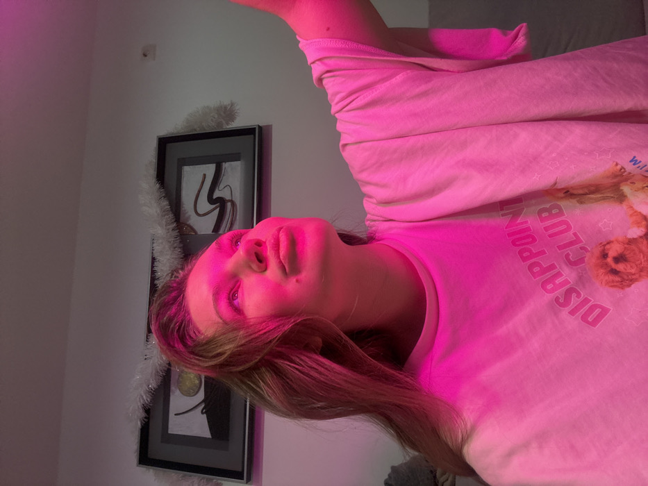 A young woman with fair skin and light brown hair is illuminated by a pink light. She wears a light-colored shirt featuring the words "DISAPPOINT CLUB" and a cartoon graphic. Behind her, a framed piece of abstract art hangs on a white wall adorned with white tinsel. The image is rotated clockwise, positioning the subject on her side. A light switch is visible on the wall.