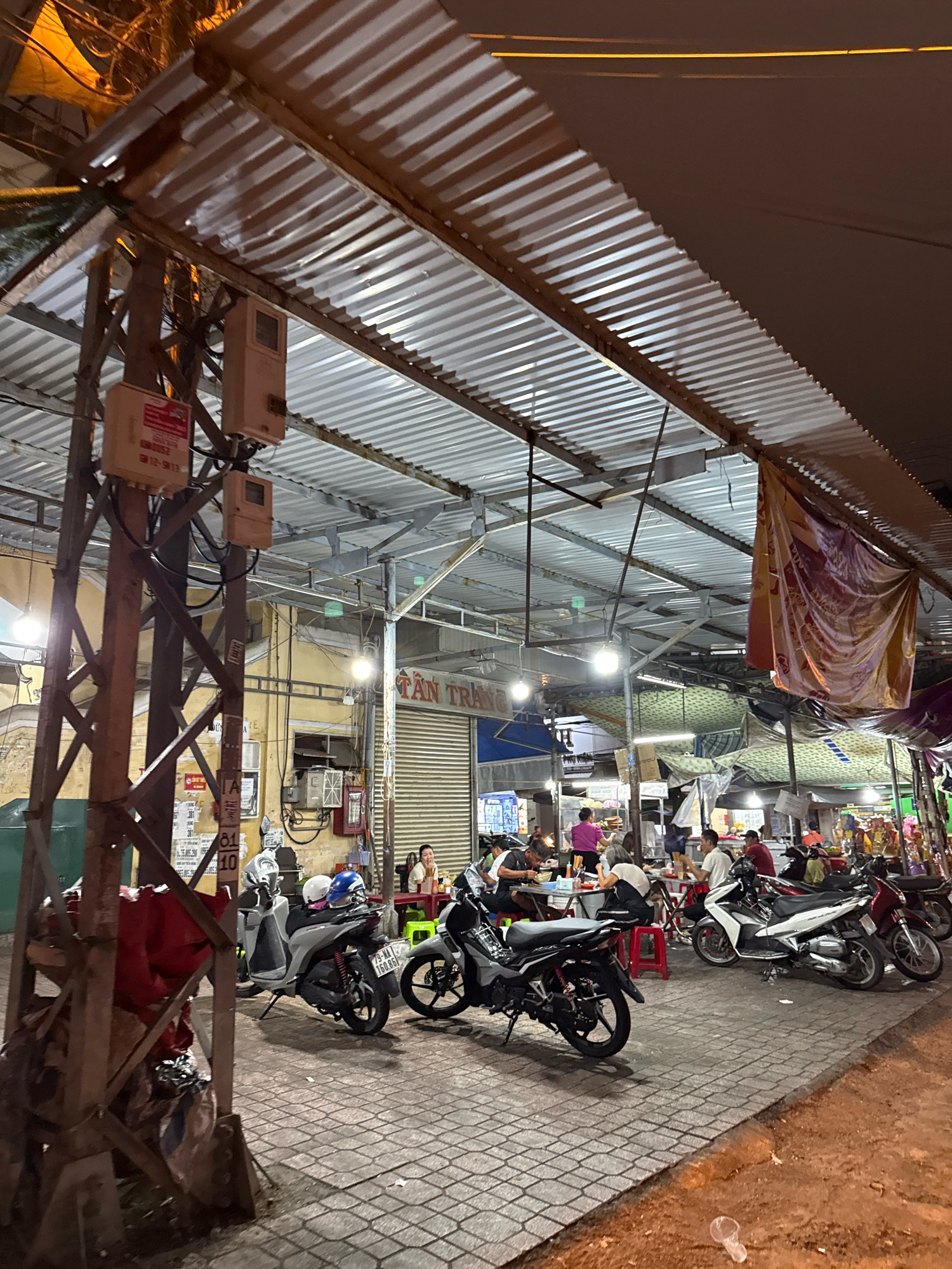 The image depicts an outdoor market or food stall area at night in Nha Trang, Vietnam. A corrugated metal roof supported by a metal frame covers the area, providing shelter. Several motorcycles are parked on the brick-paved ground. In the background, a building with yellow walls and a rolling metal door is visible, with signage that appears to read "TAN TRAN." People are seated at tables, some eating and others conversing, under the covered area. One person is standing and appears to be preparing food. Electrical boxes and wires are attached to a metal pole on the left side of the image. The lighting suggests artificial illumination from the stalls and streetlights.