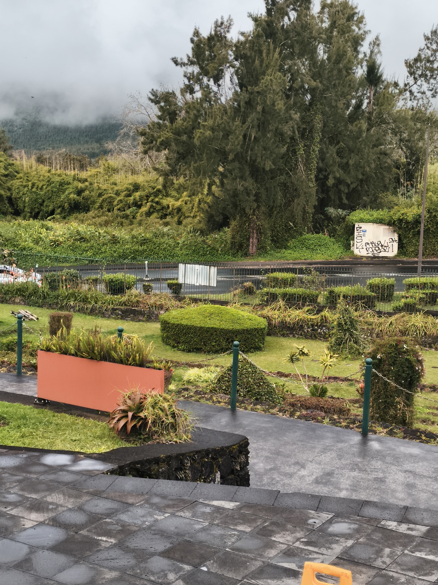 The image depicts an outdoor scene in La Plaine-des-Palmistes, France. In the foreground, a dark tiled pathway with puddles suggests recent rain. Adjacent to this is a raised planter box of an orange hue, filled with green, spiky plants. Beyond the pathway, a manicured garden area features various shrubs, including neatly trimmed hedges, and decorative plants. A low, black metal fence encloses this garden space, separating it from a road. Across the road, a white structure is visible with graffiti on its surface. In the background, dense green foliage, including bamboo and tall trees, creates a lush, natural backdrop. The sky is overcast and misty, with clouds obscuring the tops of distant, tree-covered hills. No people are visible in the image.