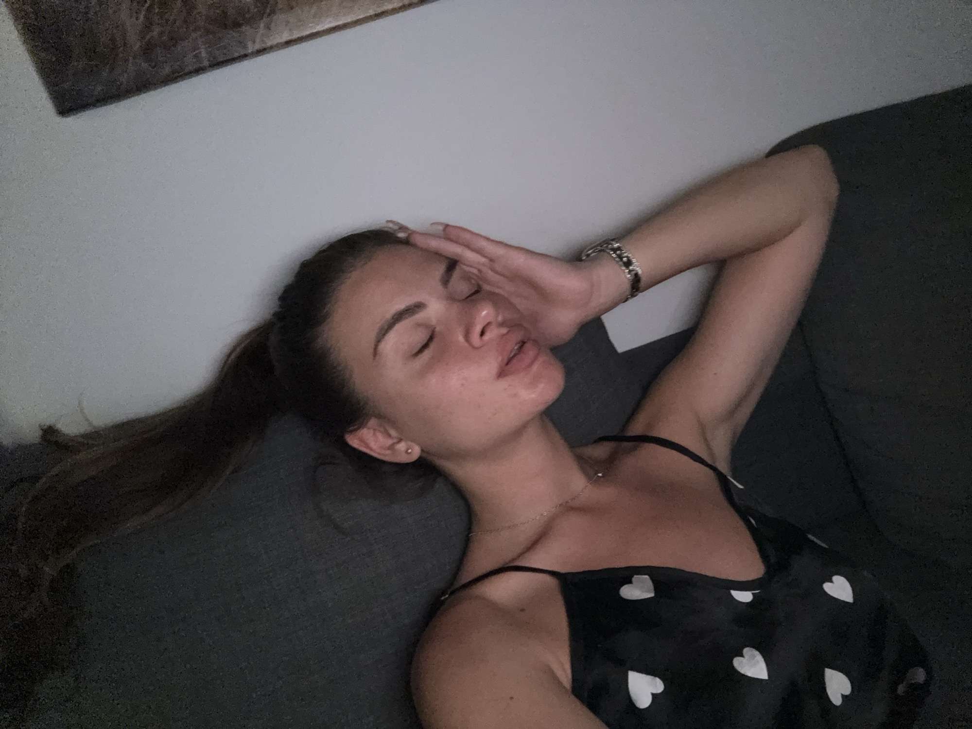 The image shows a woman lying on a dark grey couch, resting her forehead with her hand. She is wearing a black camisole with white heart patterns and a silver bracelet on her left wrist. Her eyes are closed and her mouth is slightly open. A long ponytail of dark brown hair extends to the left. A framed artwork with abstract patterns hangs on the white wall behind her. There are no visible indications of the location being in Plovdiv, Bulgaria.