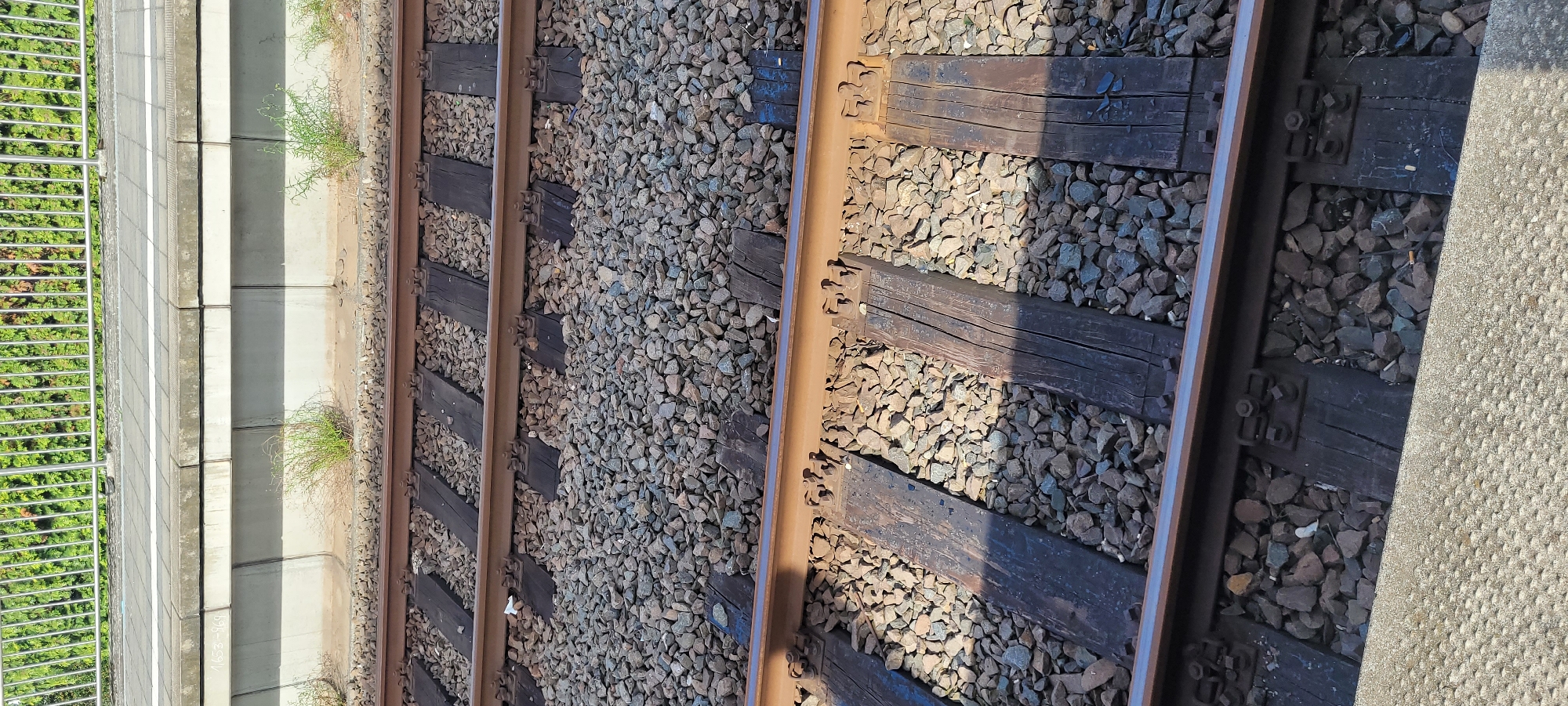 The image shows a close-up view of a set of train tracks in Rodgau, Germany. The tracks are made of steel rails supported by wooden sleepers, laid on a bed of ballast. In the background, a concrete platform with a metal railing and green foliage can be seen. The scene is outdoors, under natural lighting, with shadows cast across the tracks, indicating it is daytime. There are no people or vehicles visible in the image.