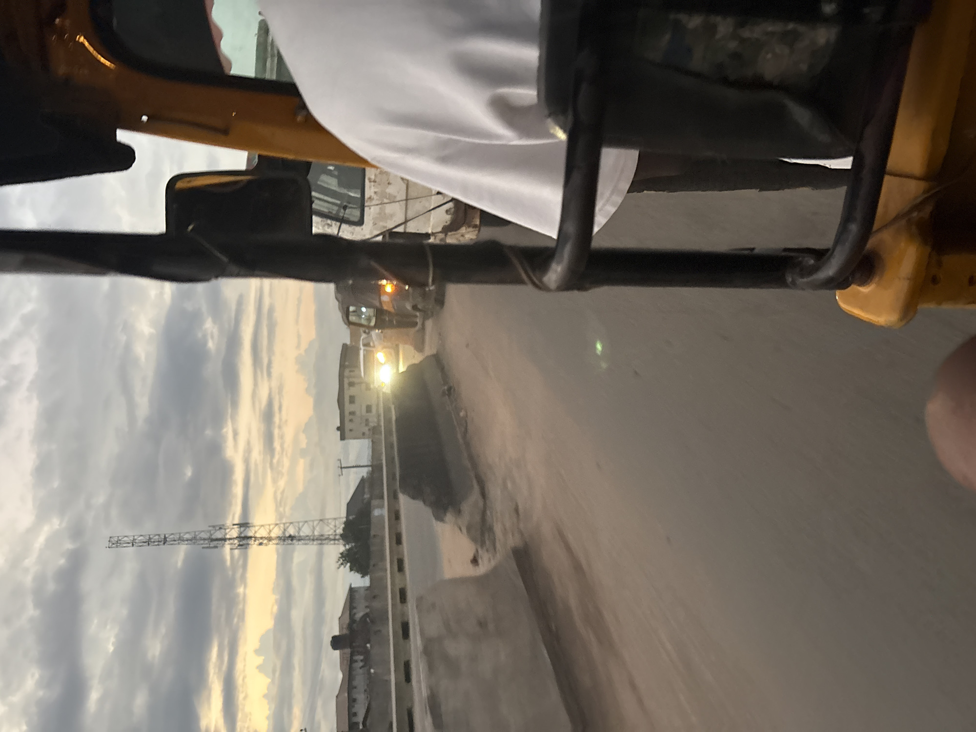 The image depicts a street scene in Onitsha, Nigeria, likely taken from a moving vehicle, possibly an auto-rickshaw, given the visible frame and handlebars. In the foreground, a person wearing a white t-shirt is partially visible, their back turned towards the camera. The vehicle's yellow frame and black metal bars are prominent.
The road ahead appears to be in a state of disrepair, with concrete barriers that are partially broken. Several vehicles are visible on the road, including a dark auto-rickshaw with its taillights illuminated, and a white van. The headlights of an oncoming vehicle are bright, suggesting dusk or a cloudy day.
In the background, a tall telecommunications tower stands against a cloudy sky with hints of sunset colors. Buildings, some appearing residential, are visible along the roadside. The overall environment suggests a bustling urban setting in Nigeria.
