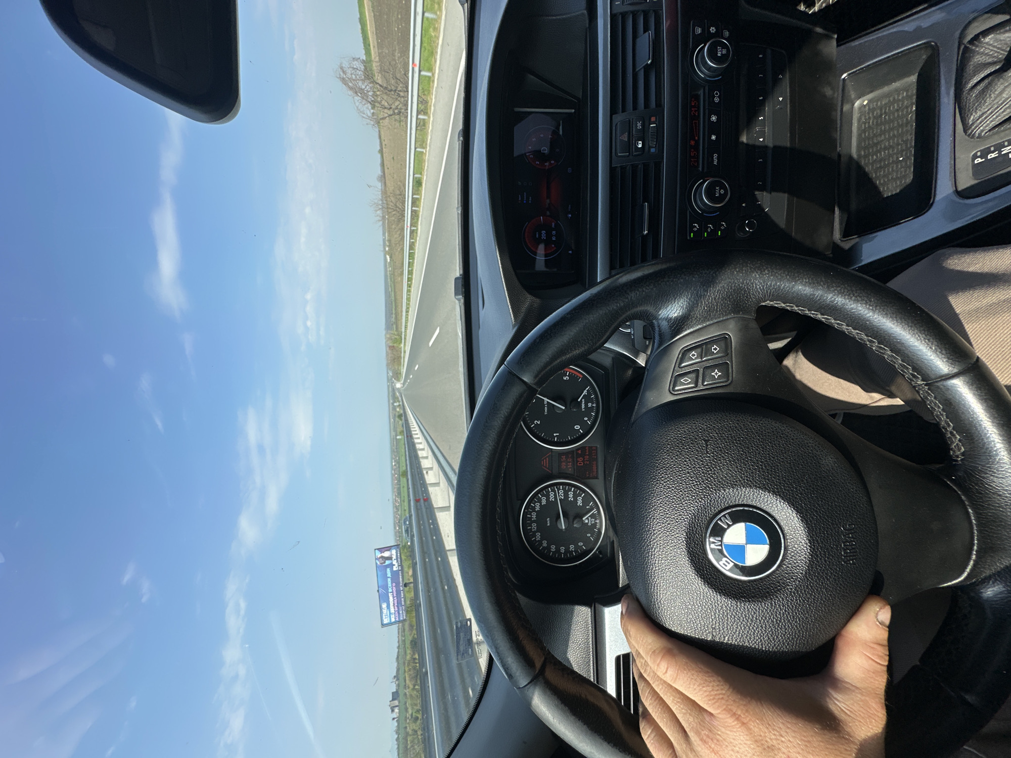 The image depicts the interior of a BMW vehicle, specifically the driver's perspective. The steering wheel, with the BMW logo prominently displayed, is the central object. The driver's hand is visible holding the steering wheel. The dashboard, including the speedometer and other gauges, is in focus. The car is traveling on a highway, as evidenced by the road visible through the windshield. A roadside billboard displaying the text "NETTHALAD" is on the right side. The sky is clear and blue. Based on the identified text on the billboard, the location is Rupkite, Bulgaria. The car is showing a mileage of 213.9 kilometers. The temperature is showing 21.5 degrees Celsius. The speedometer indicates a speed of 209 kilometers per hour.