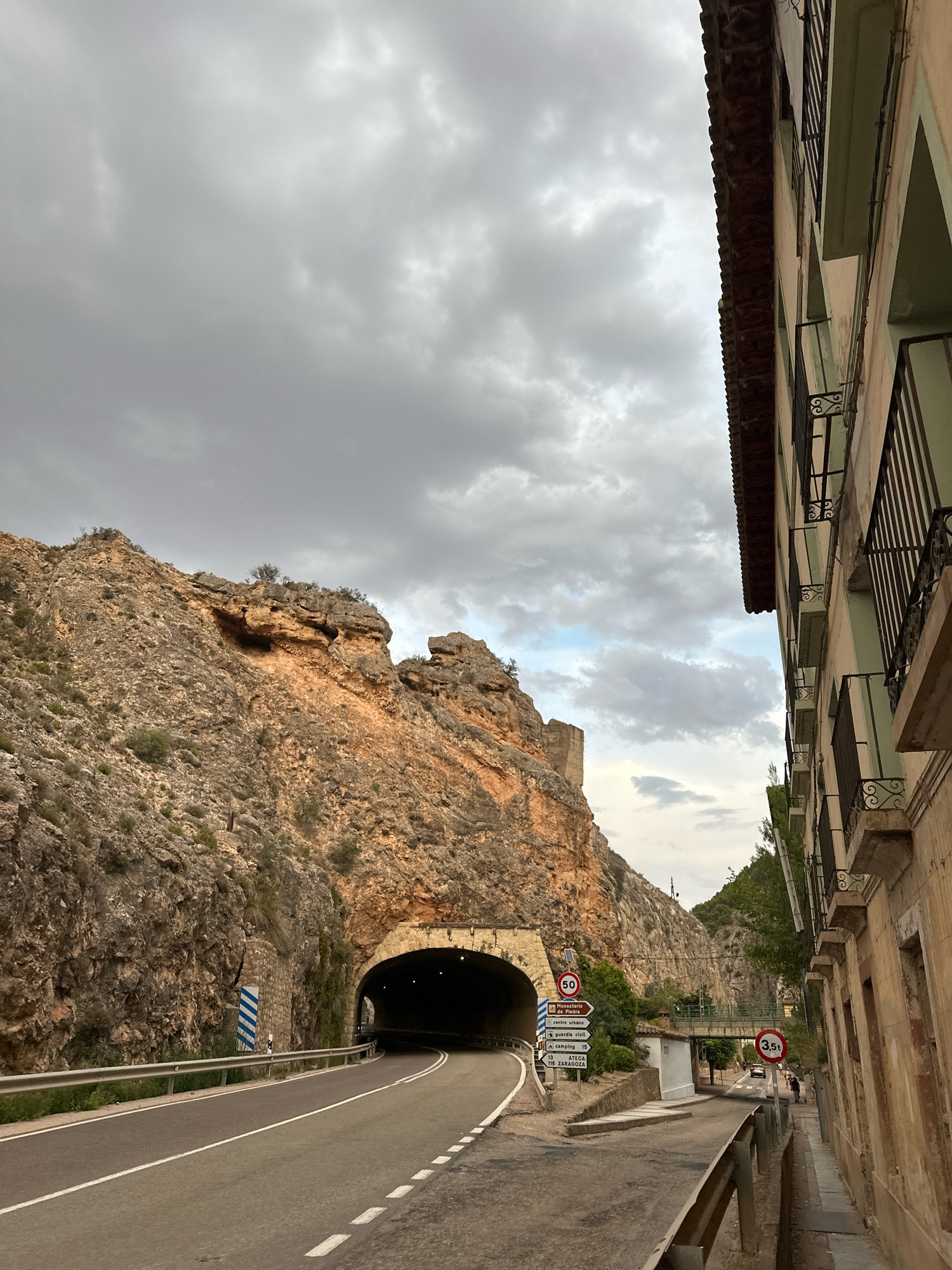 This is a horizontal, eye-level, outdoor photograph of a road leading into a tunnel through a rocky mountainside. On the right is the side of a weathered, multi-story building with balconies. The sky is overcast with large, grey clouds. The road curves to the left as it enters the tunnel. There are signs on the side of the road. One sign indicates a speed limit of 50. Another sign shows directions to "Monasterio de Piedra", "centro urbano", "guardia civil", "camping", and "A15 ZARAGOZA." Another circular sign indicates a price of 3.5€. A person is visible on the sidewalk near the signs.
