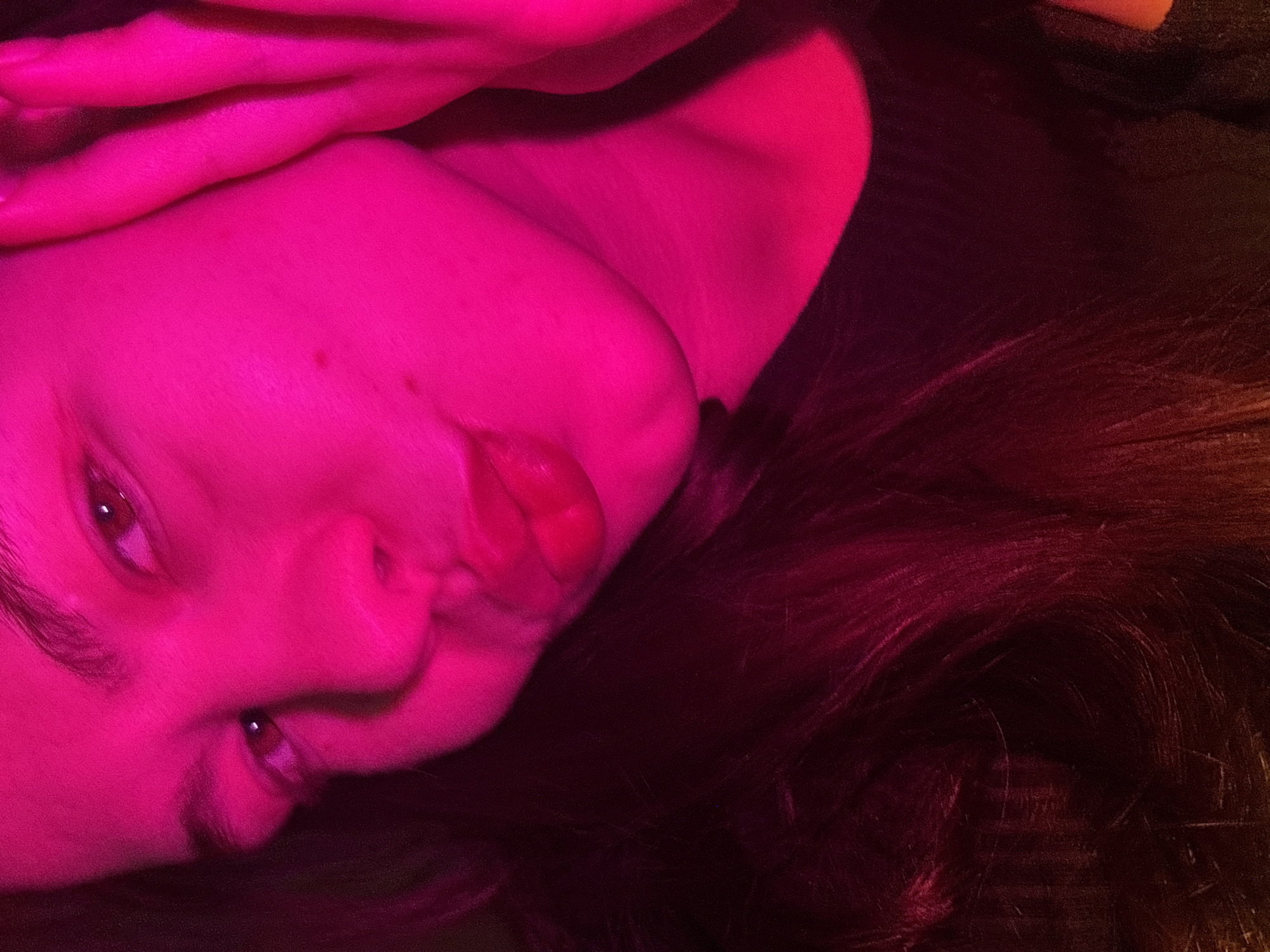 The image depicts a close-up of a person's face and shoulder in a predominantly pink lighting environment. The person's facial features are visible, including their eyes, nose, and lips. Their hair is dark and visible on the right side of the frame. The skin appears smooth. No other details about the person, their actions, interactions, or the environment are discernible. No location is specified.